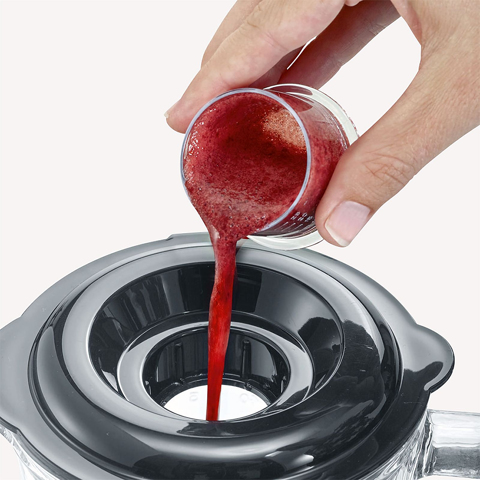 A hand pouring red liquid from a measuring cup into a blender.