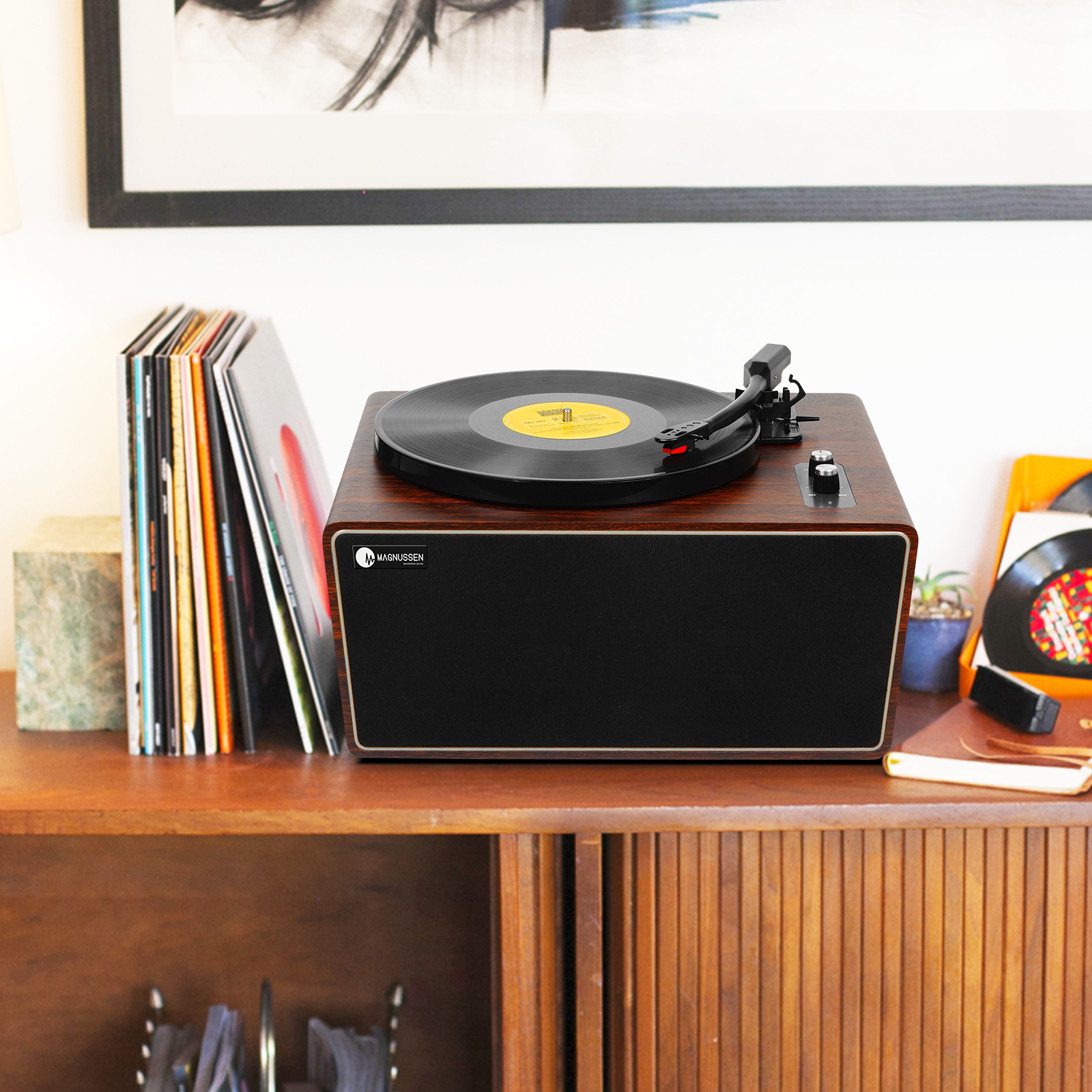 Tocadiscos sobre un mueble de madera con discos y una planta. Hay un cuadro colgado en la pared.