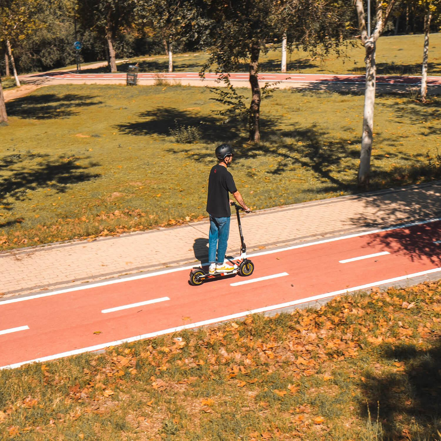 Un hombre montando un scooter en un camino rojo. El fondo es un parque.
