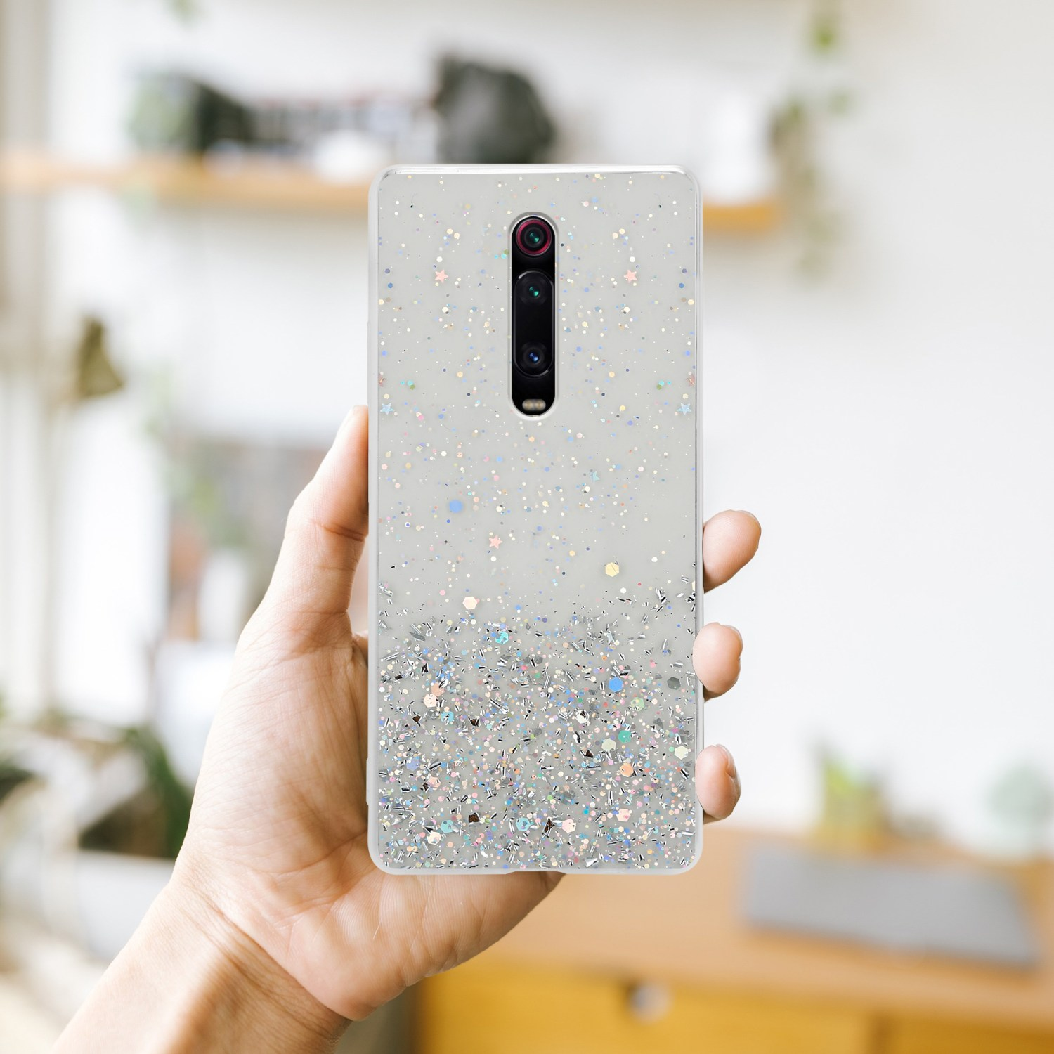 Mano che tiene una custodia per telefono con design glitterato, sfondo sfocato con una stanza.