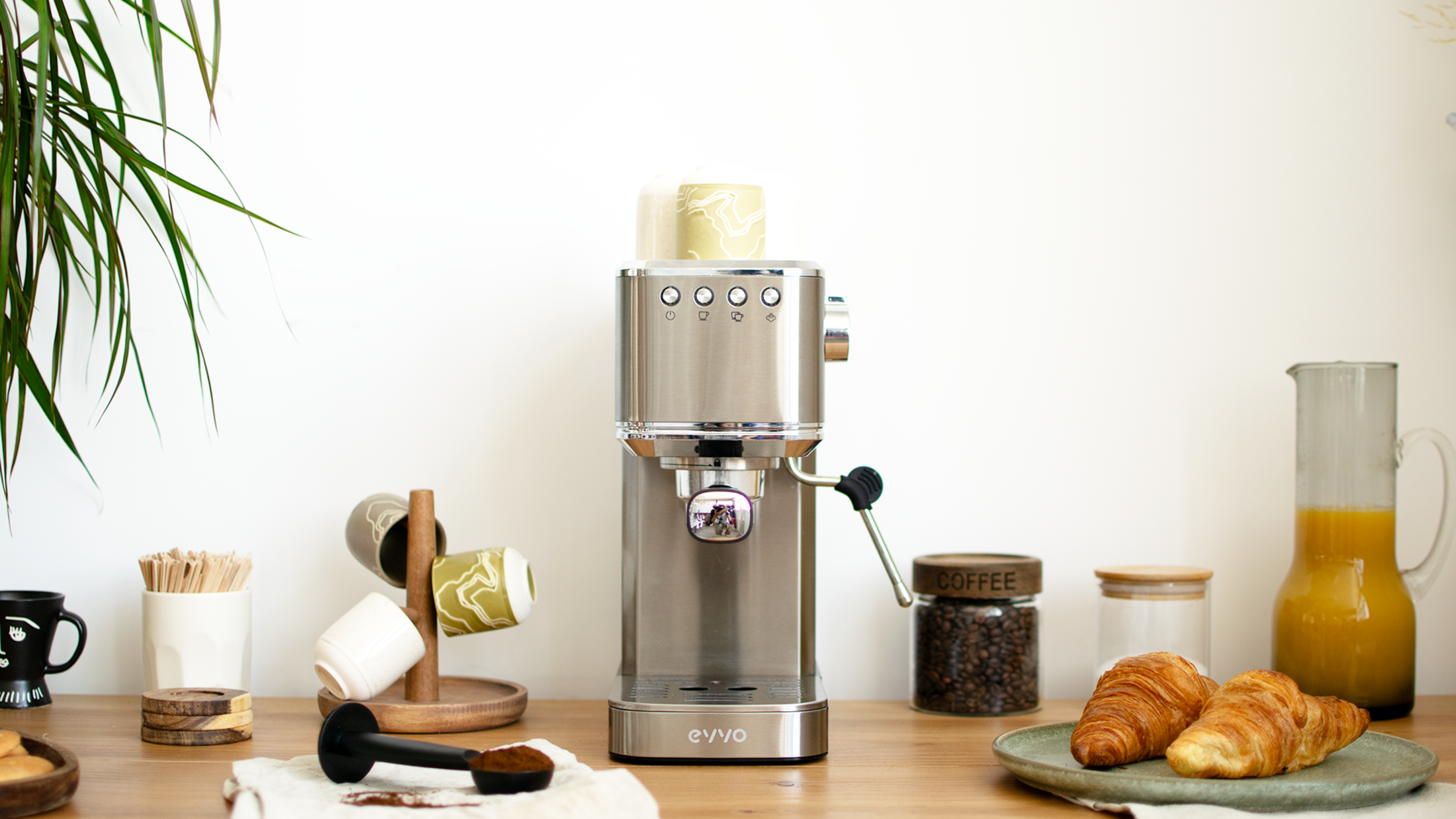 Cafetera espresso de acero inoxidable sobre una mesa de madera. Granos de café, cruasanes y otros accesorios presentes.