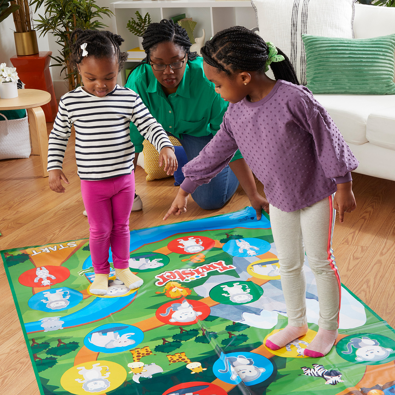 Zwei junge Mädchen und eine Frau spielen Twister auf einer Matte mit bunten Kreisen und Tierformen.