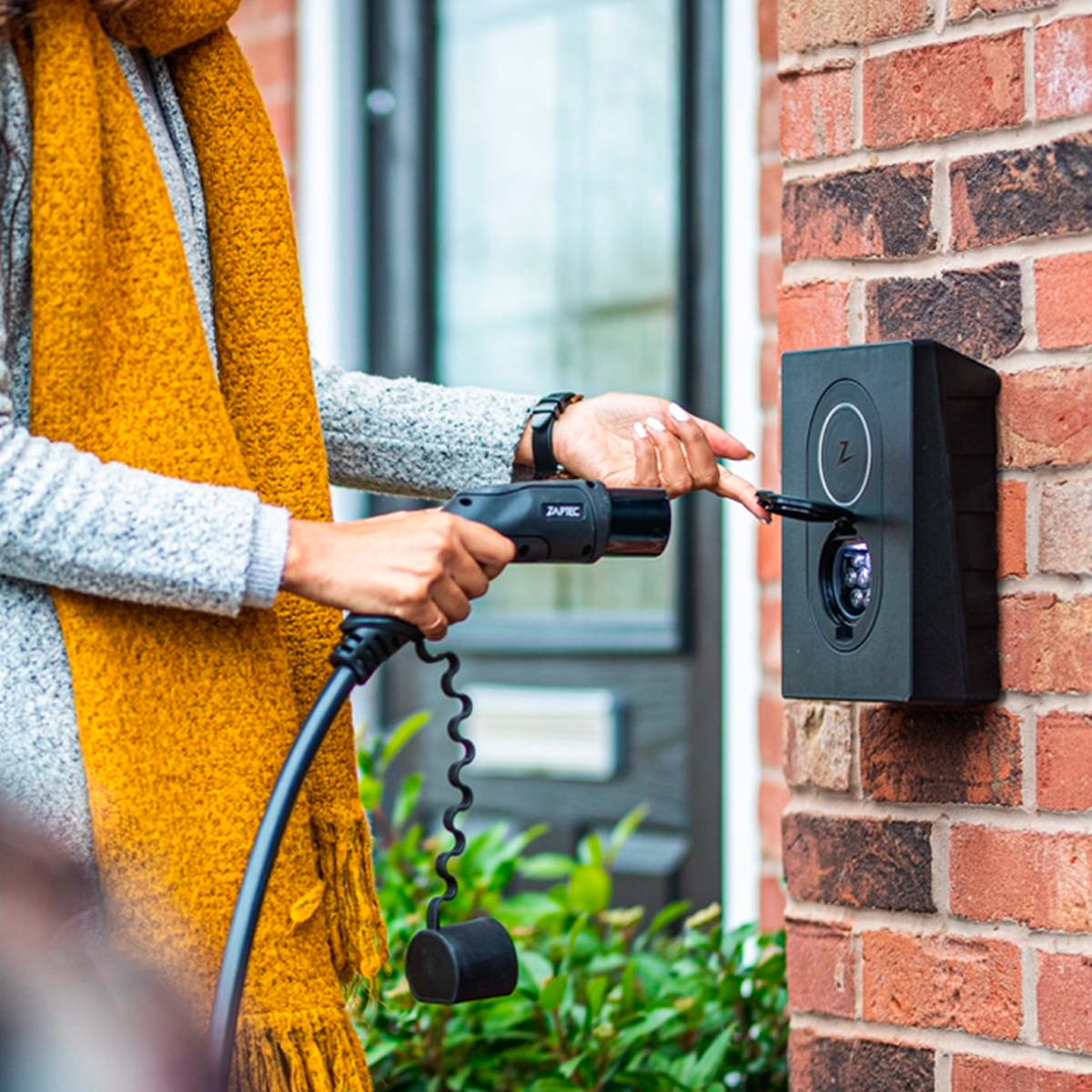 A person is using a ZAPTEC charger on a brick wall. A yellow scarf is worn.