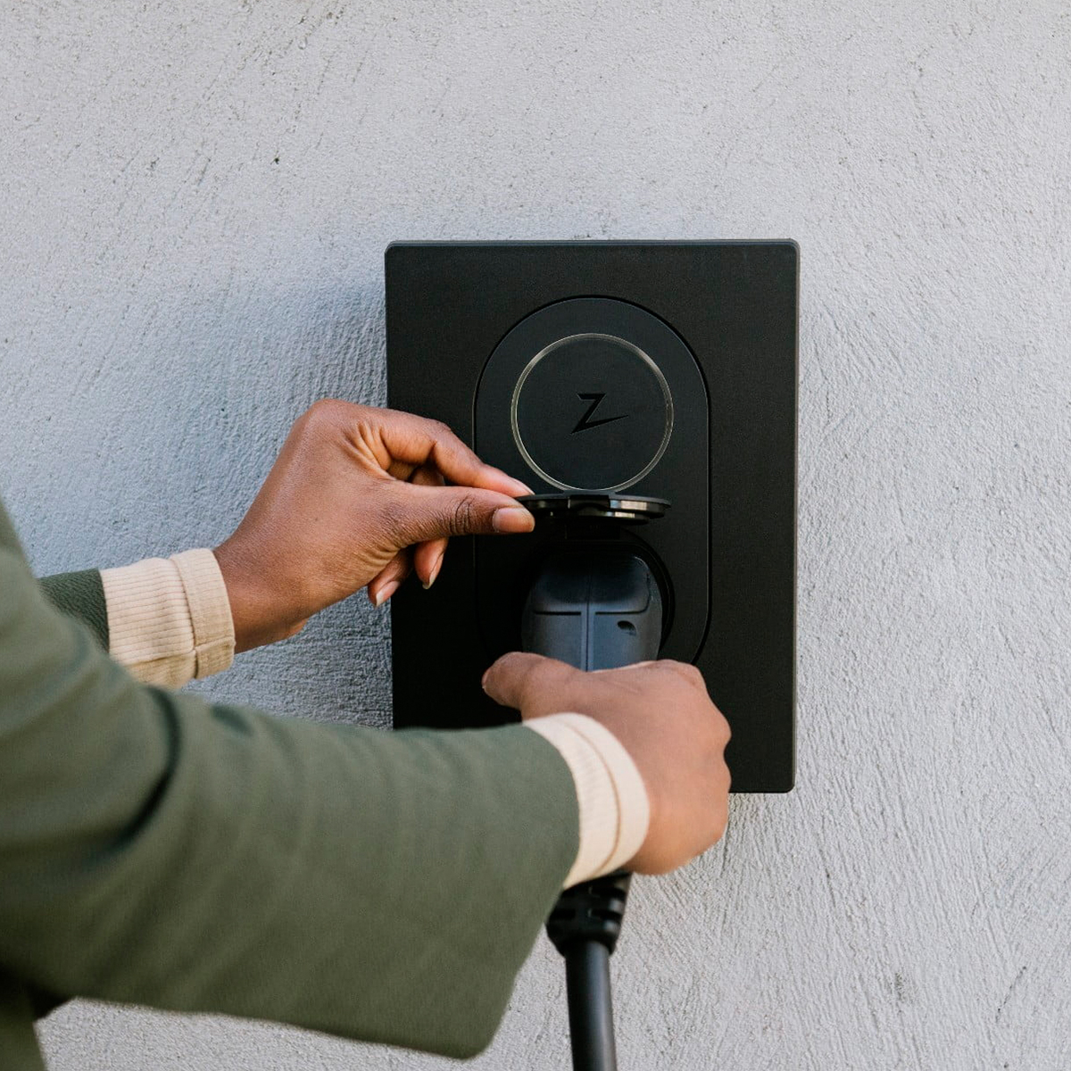 Hands plug a charger into an EV charging station on a gray wall.