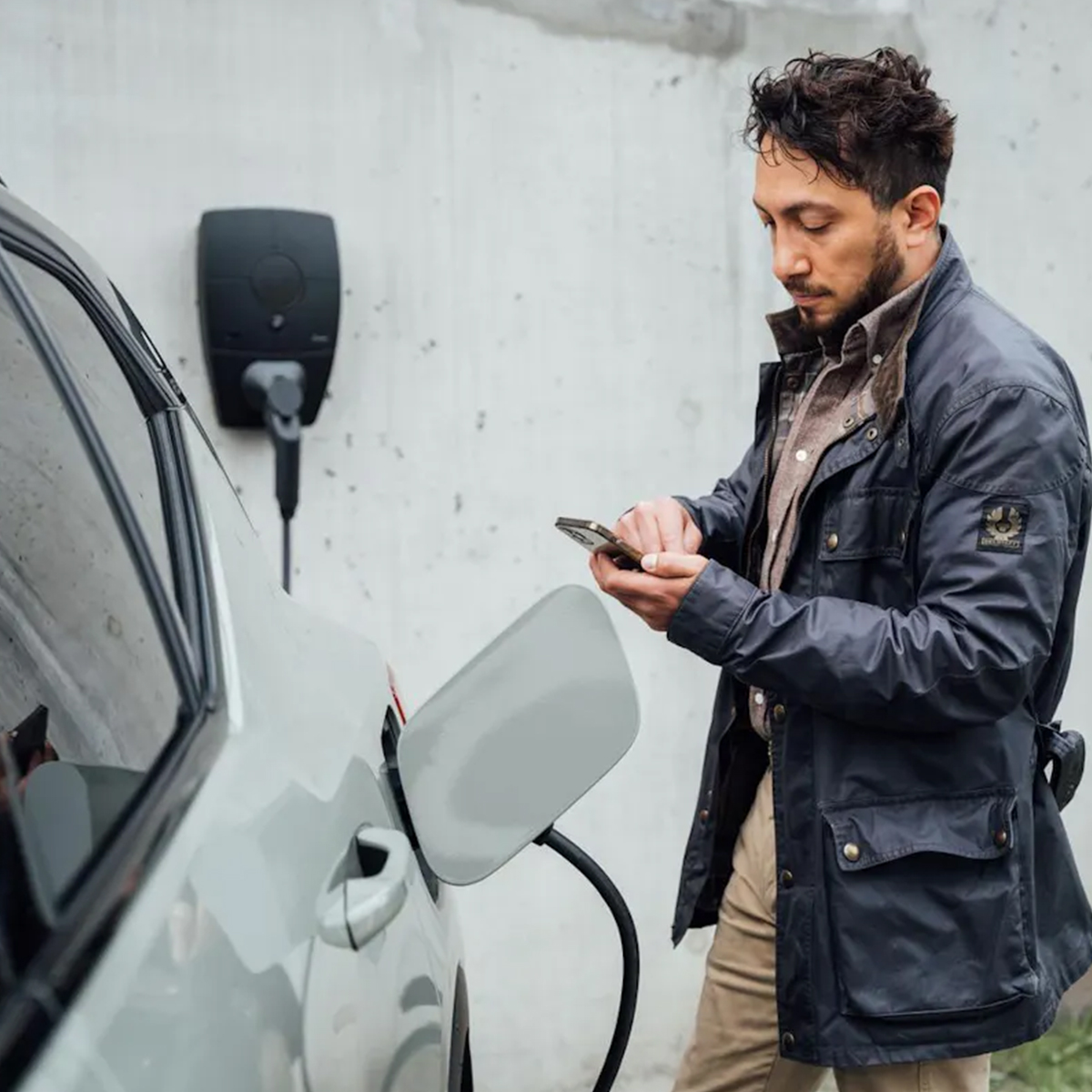 Man by a car charging. He's holding a phone. The car is being charged.