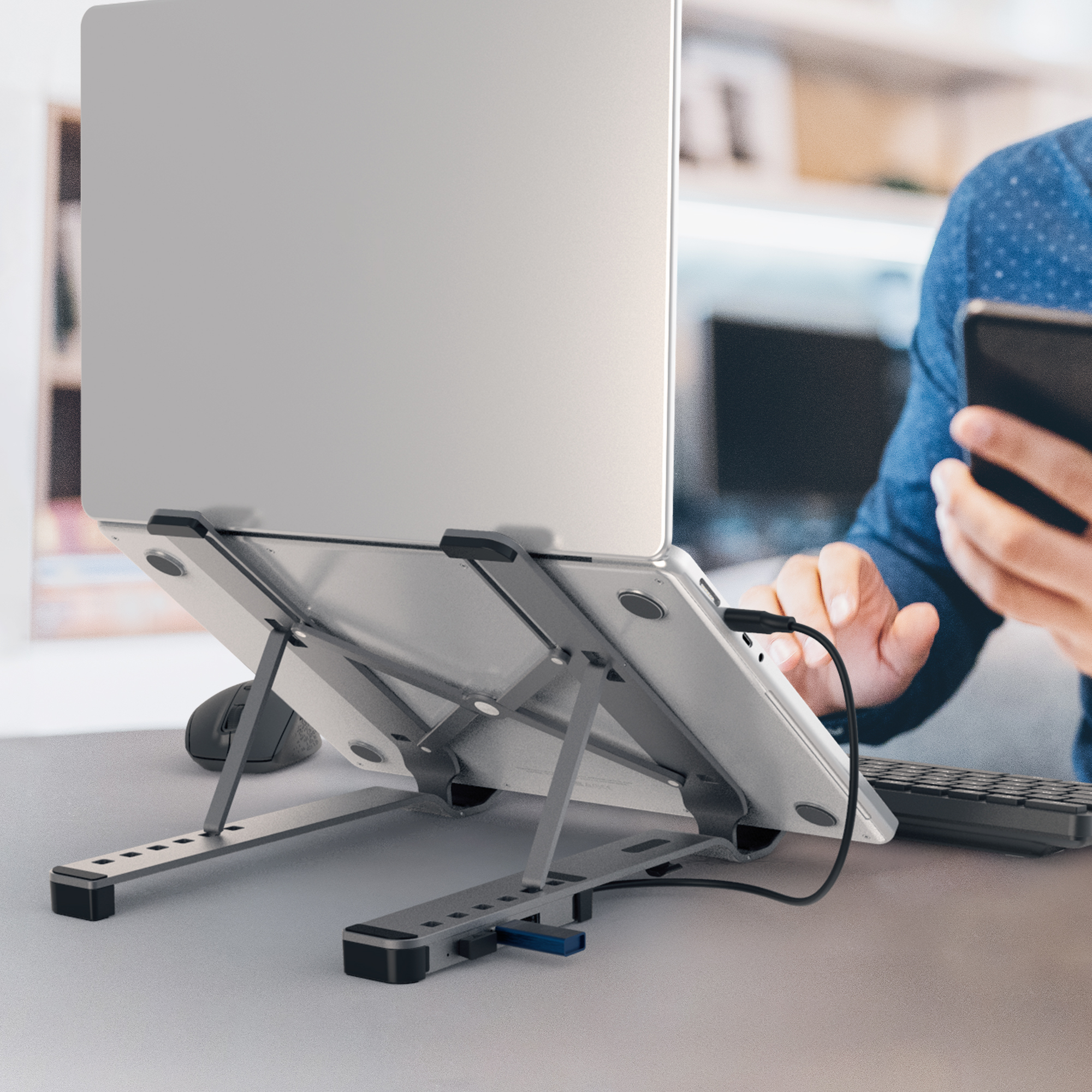Ein Laptop auf einem silbernen Ständer, mit Tastatur und Maus. Eine Person benutzt ein Telefon.
