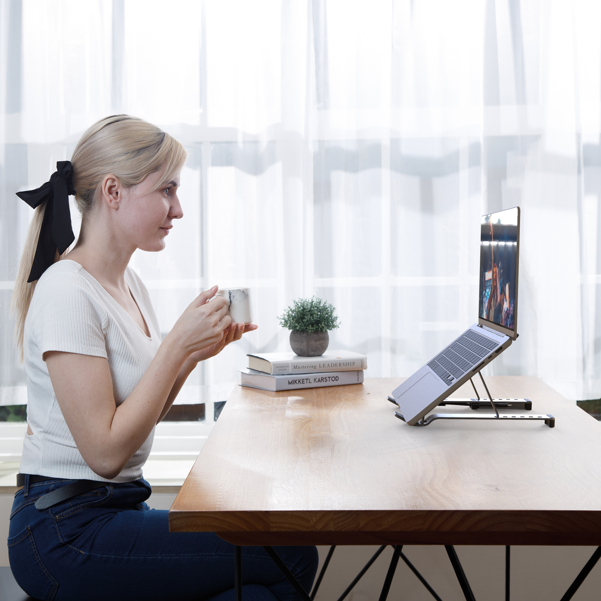 Frau benutzt Laptop auf Ständer, hält Tasse. Bücher auf dem Tisch, Vorhänge im Hintergrund.