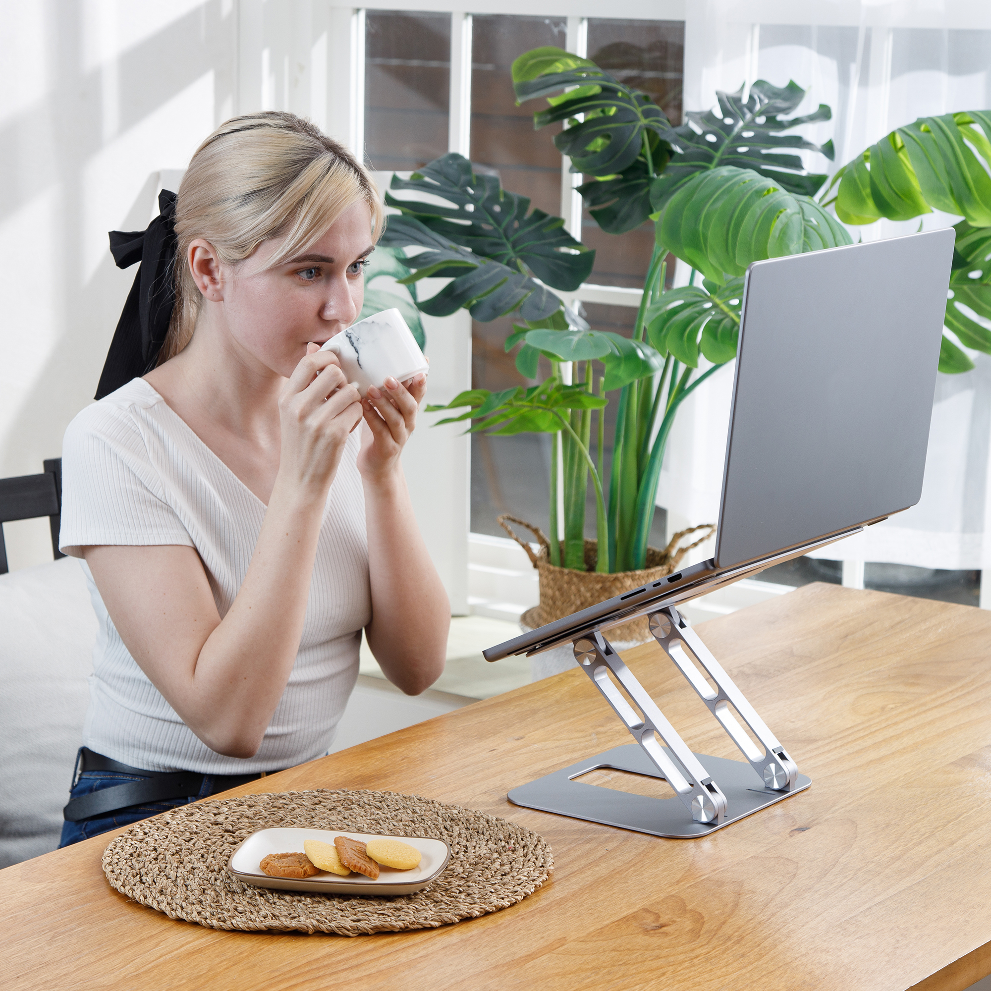 Woman drinking coffee, using laptop on a stand. Cookies and plant on the table.