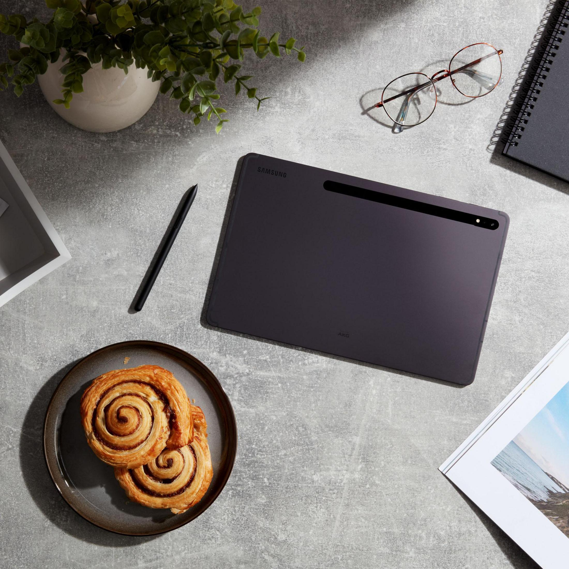 Draufsicht auf ein Tablet, einen Stift, einen Teller mit Gebäck, eine Brille und ein Notizbuch.