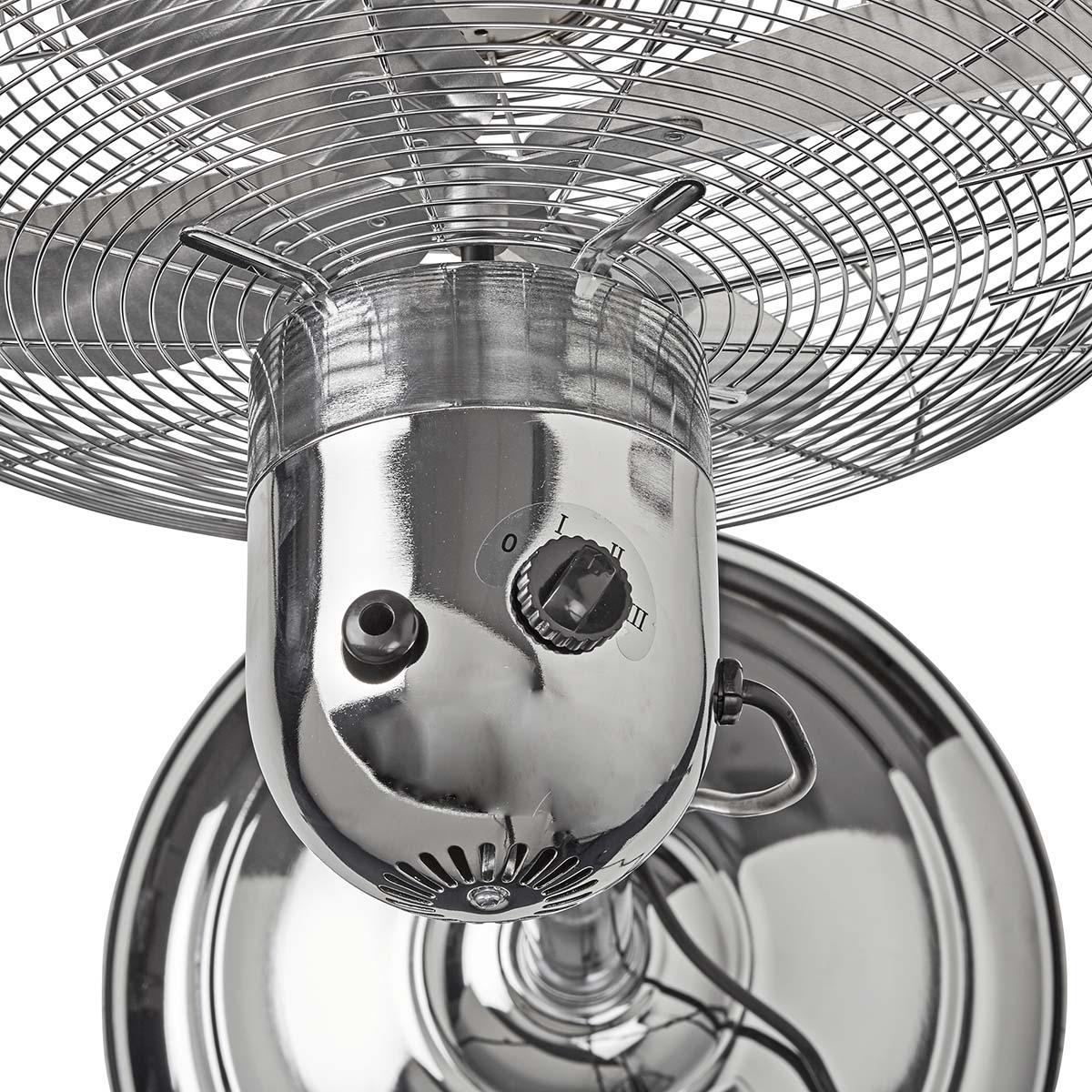 Un ventilatore cromato con un quadrante e una gabbia. La ventola è da un'angolazione bassa.