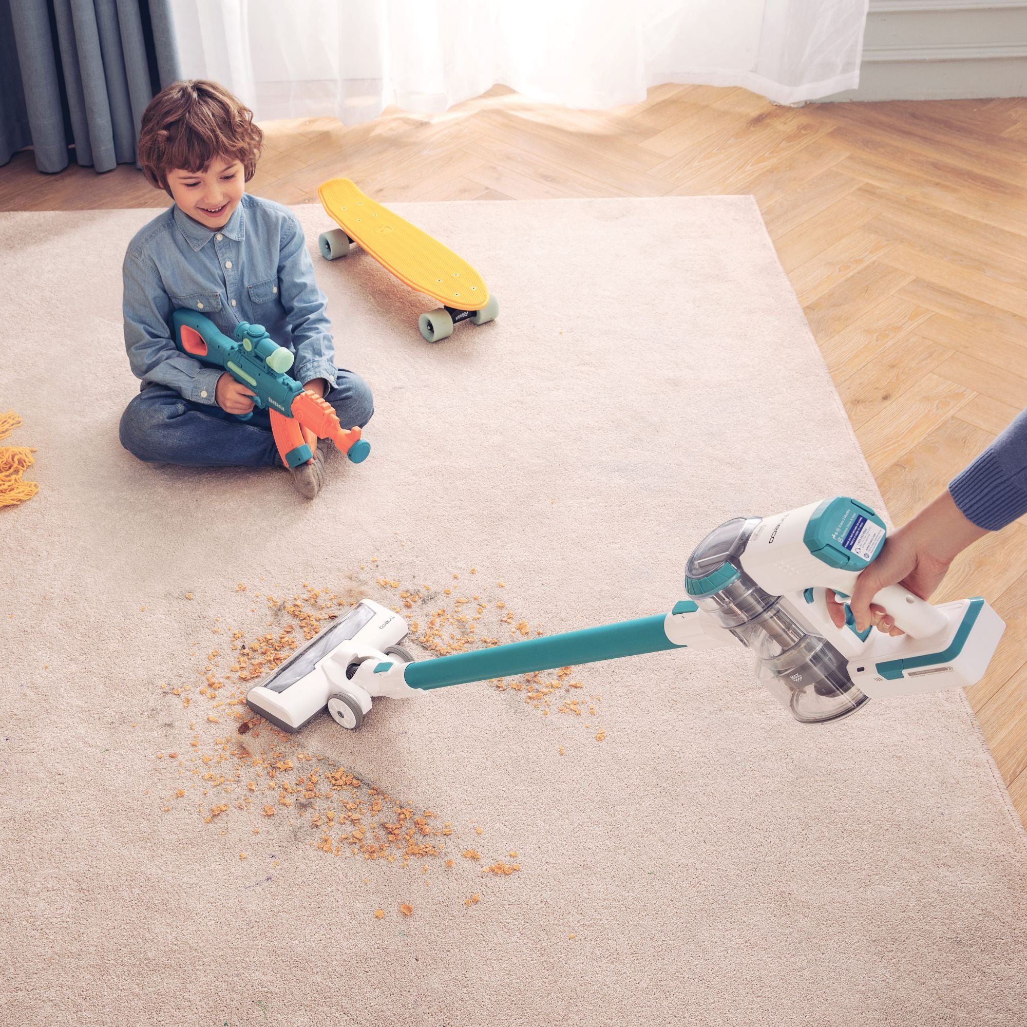 Un garçon jouant avec un pistolet jouet et un aspirateur sur un tapis. Une personne tient l'aspirateur.