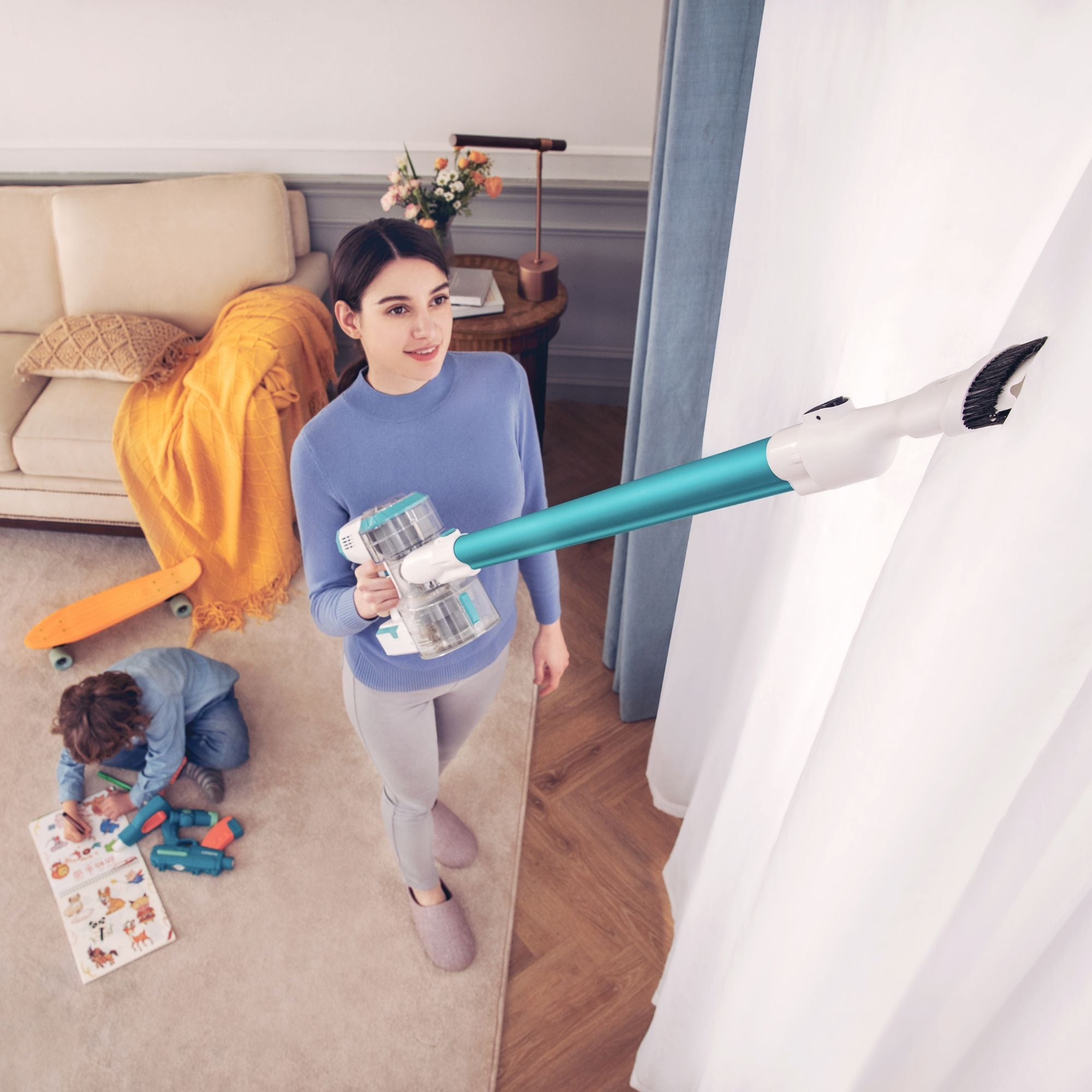 Femme utilisant un aspirateur sans fil. Enfant jouant au sol. Prise de vue intérieure avec canapé et fenêtre.