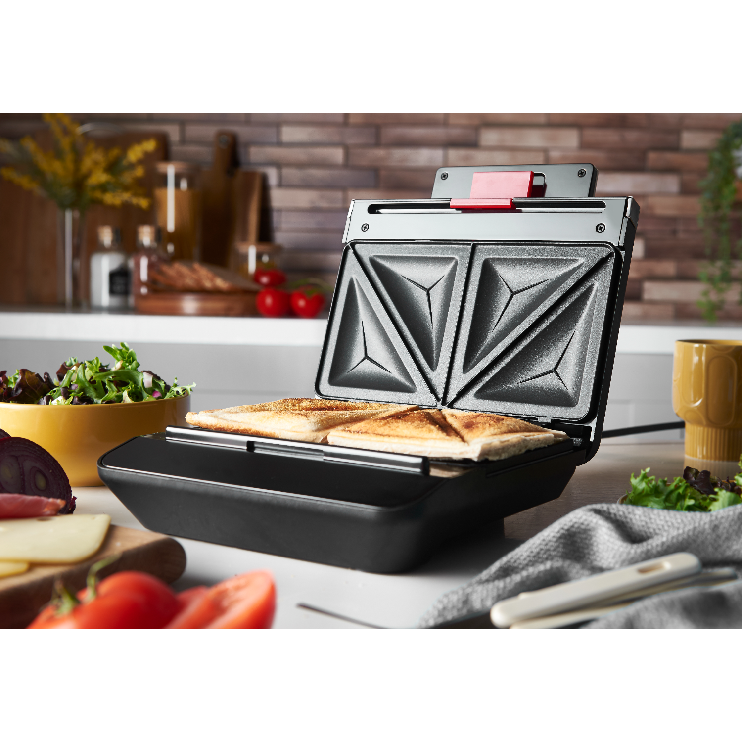 A sandwich maker with two toasted sandwiches and ingredients on the counter.
