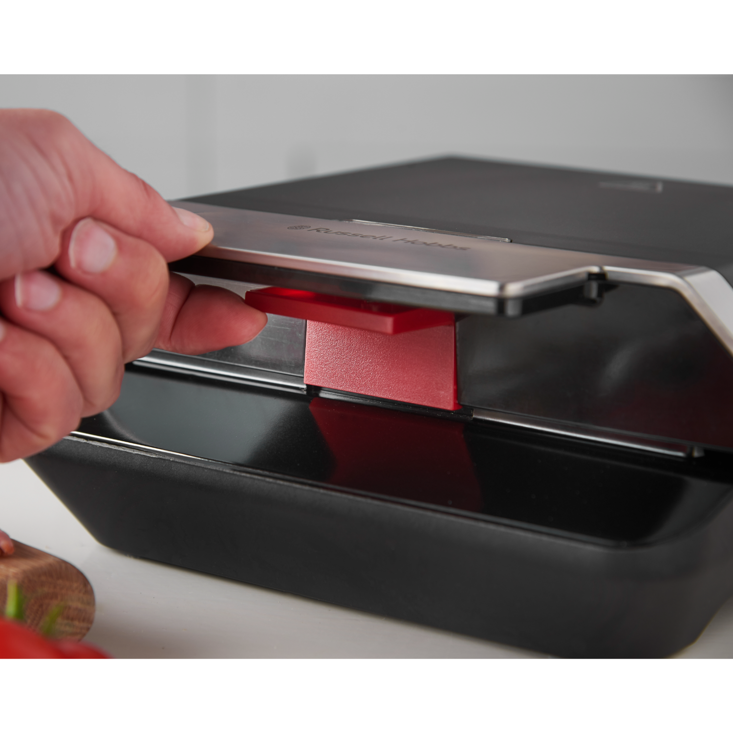 A hand opening the lid of a black sandwich maker with a red plate inside.