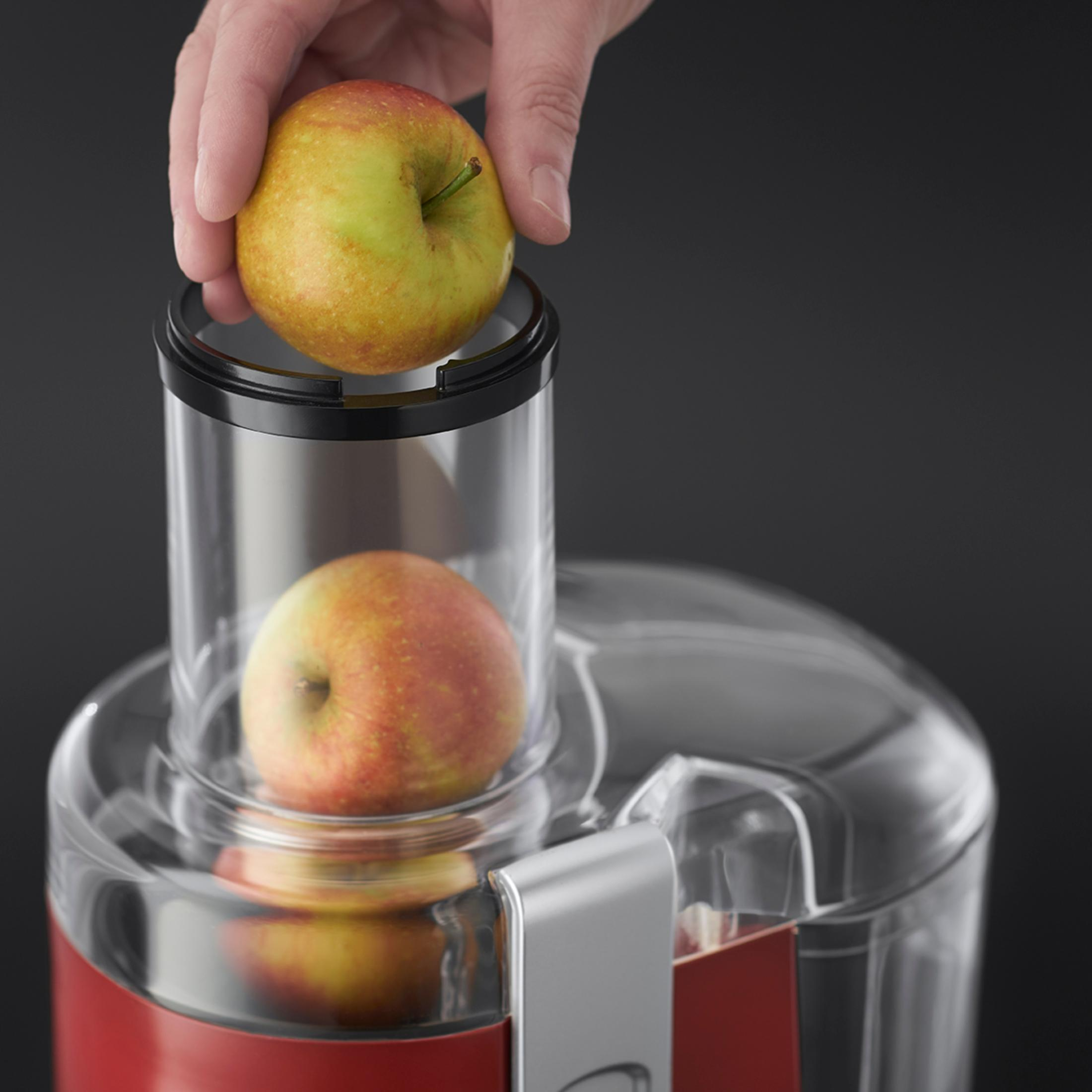 Una mano che inserisce una mela nella parte superiore di un estrattore di succo rosso e trasparente, con un'altra mela all'interno.