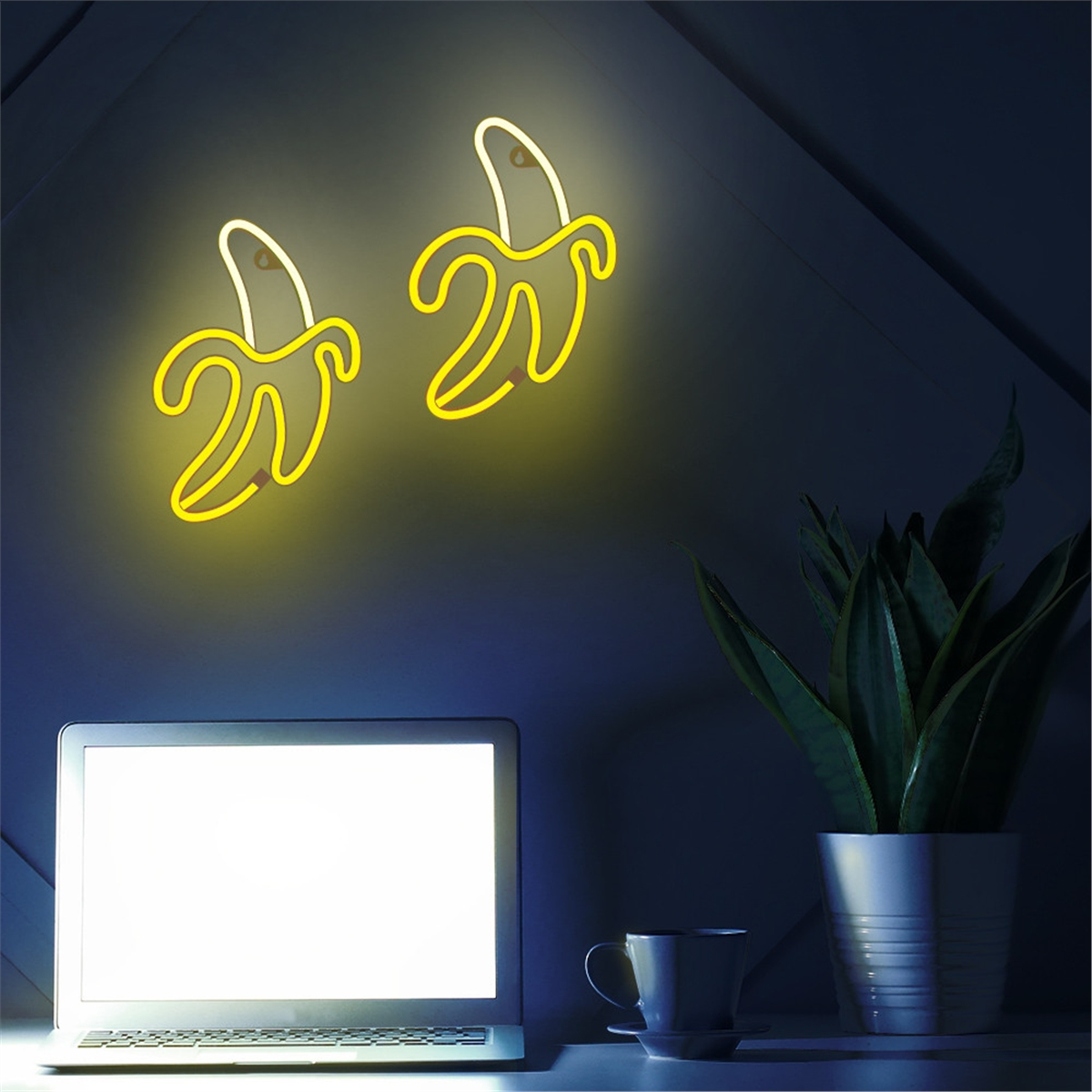 Zwei Neon-Bananenlichter an der Wand. Ein Laptop, eine Tasse und eine Pflanze sind auf einem Schreibtisch sichtbar.