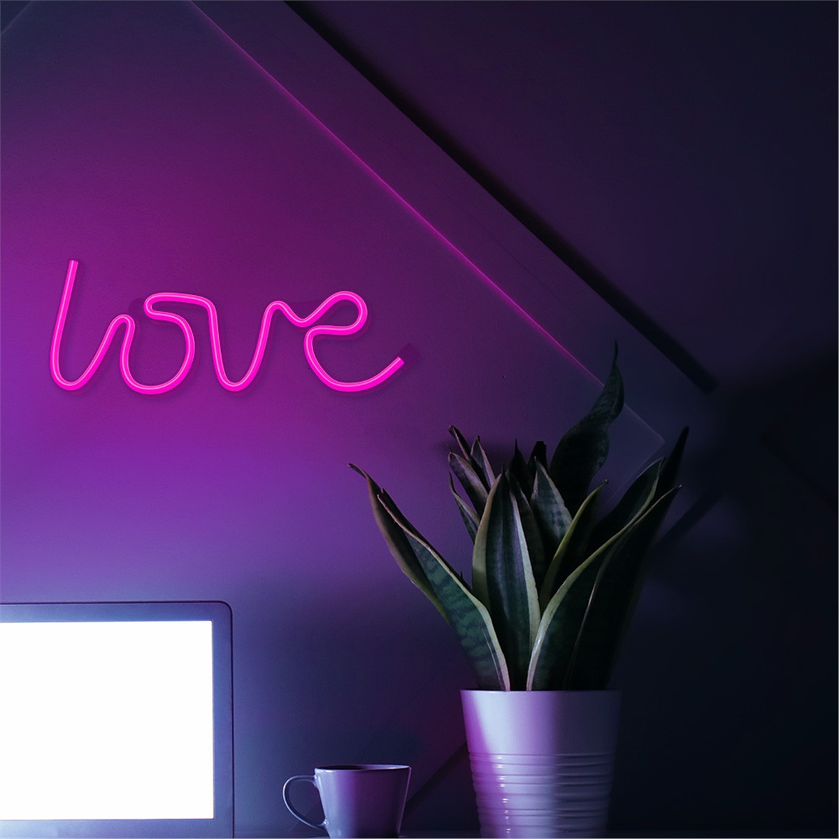 Un letrero rosa neón de 'amor' en una pared, con una planta, una taza y un portátil en primer plano.