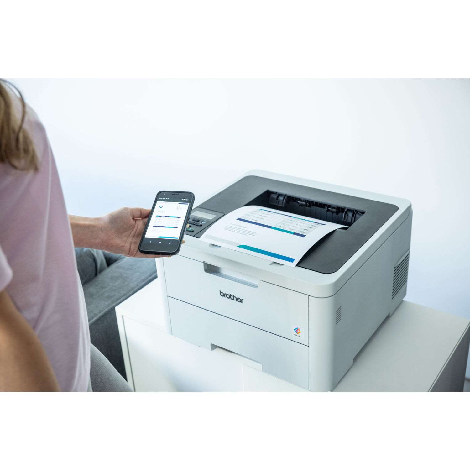 Kobieta trzyma smartfona obok białej drukarki. Z drukarki wychodzi kartka papieru.