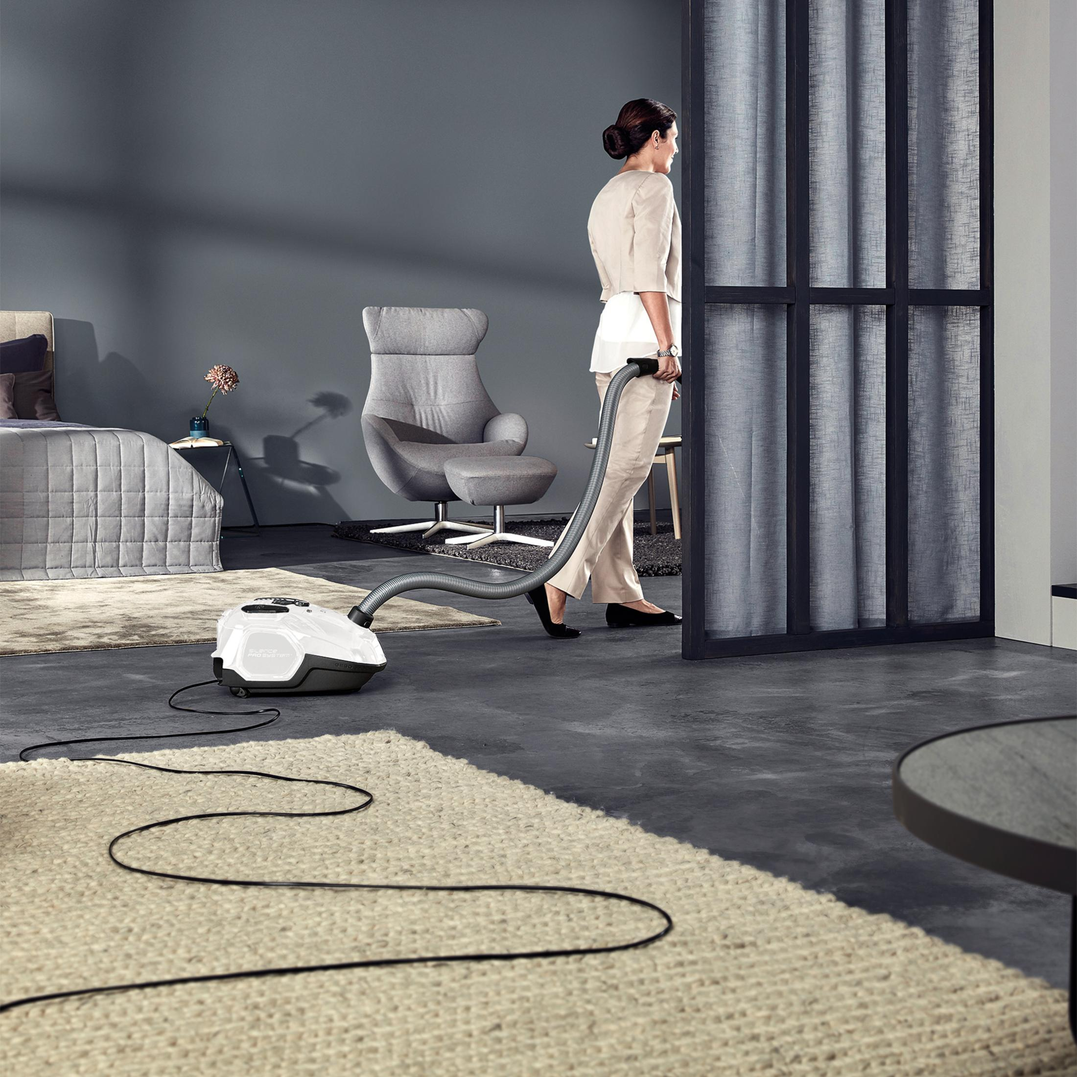 Kobieta odkurza pokój z szarymi ścianami, dywanem i szarym fotelem. Odkurzacz na pierwszym planie.