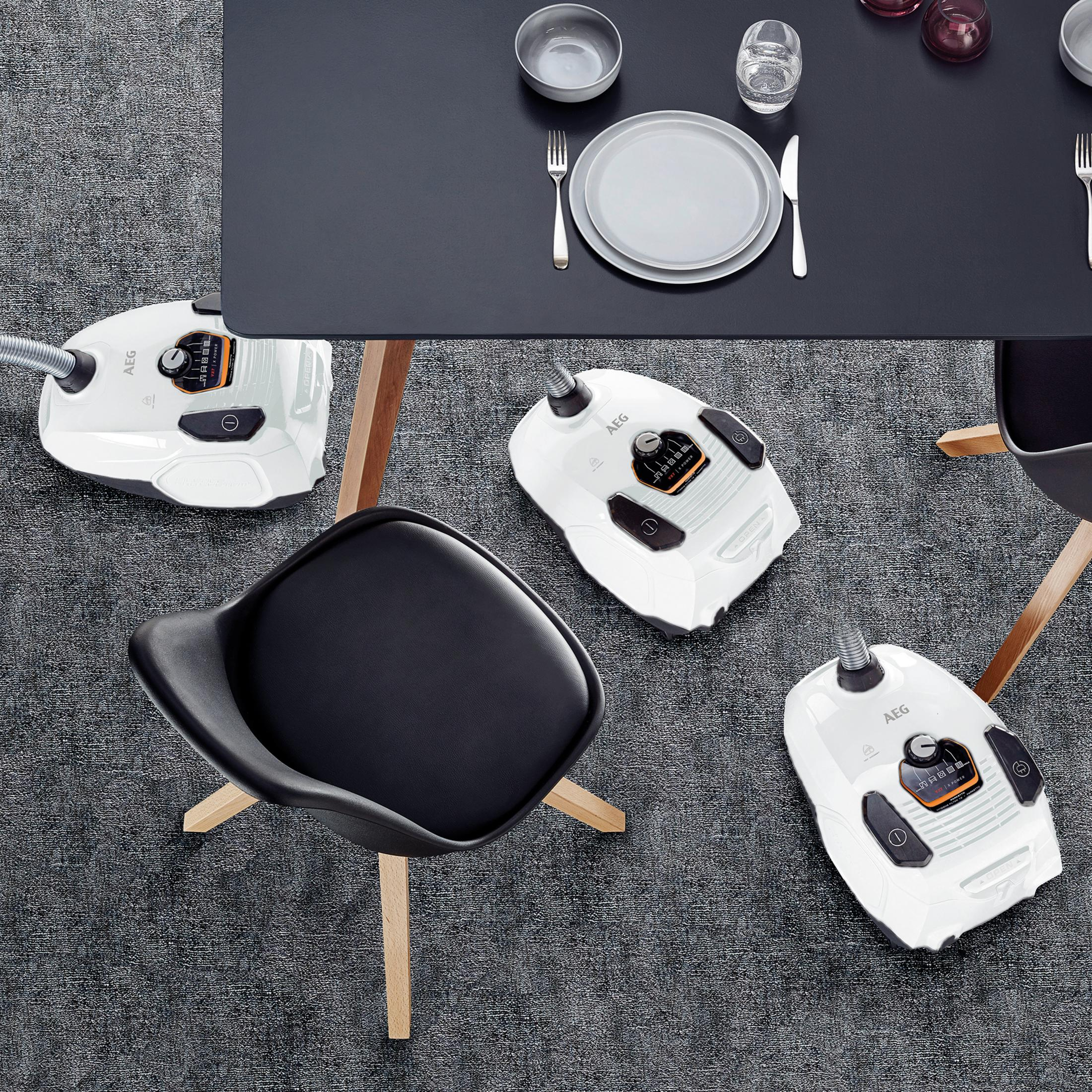 Trzy białe odkurzacze pod czarnym stołem, szary dywan, czarne krzesło i sztućce.