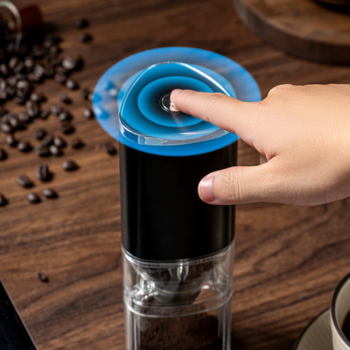 Hand drückt den Knopf auf einer schwarzen und durchsichtigen Kaffeemühle, Kaffeebohnen sichtbar.