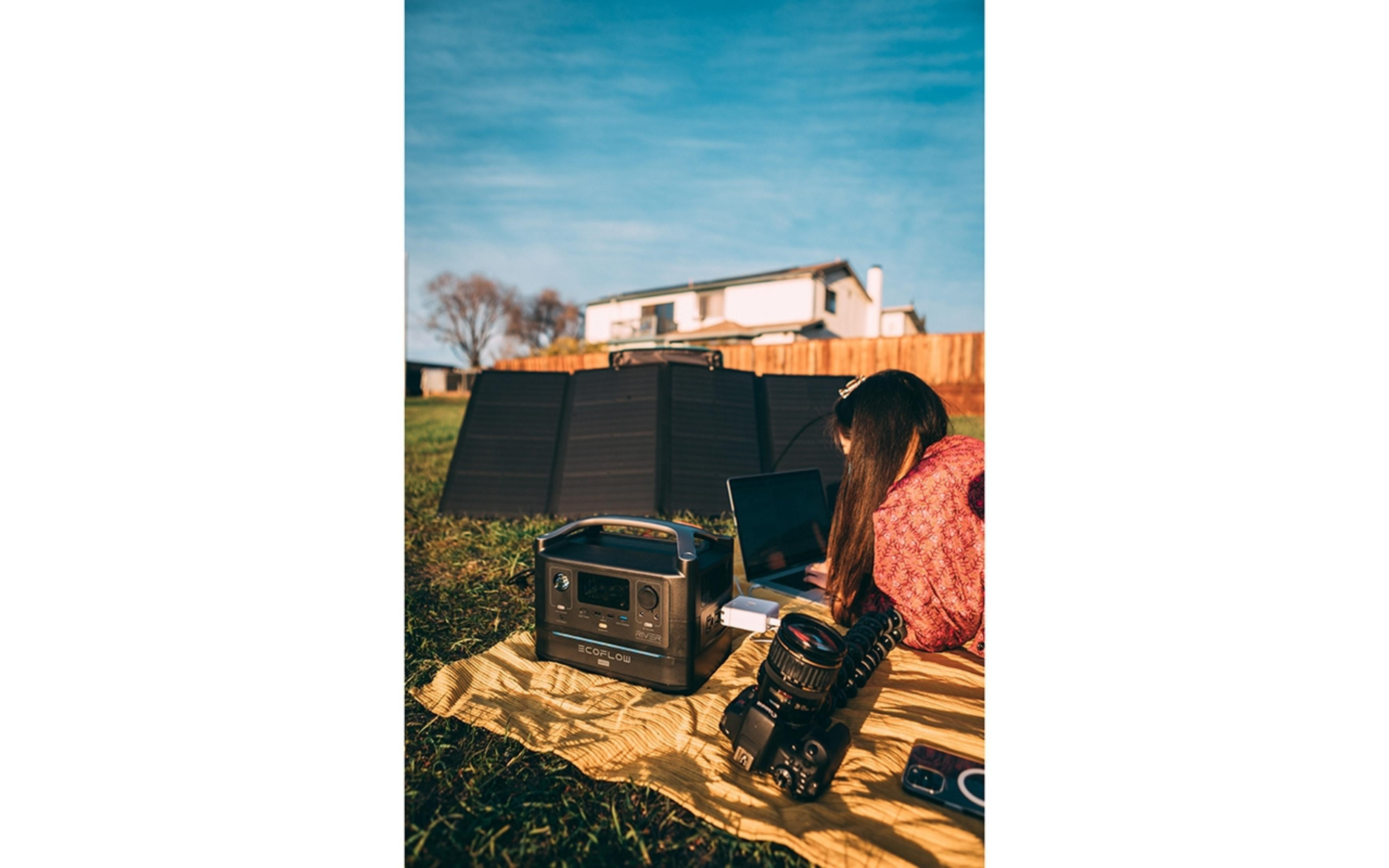 Kobieta pracuje na laptopie, panele słoneczne i elektrownia na trawie, słoneczny dzień, dom w tle.