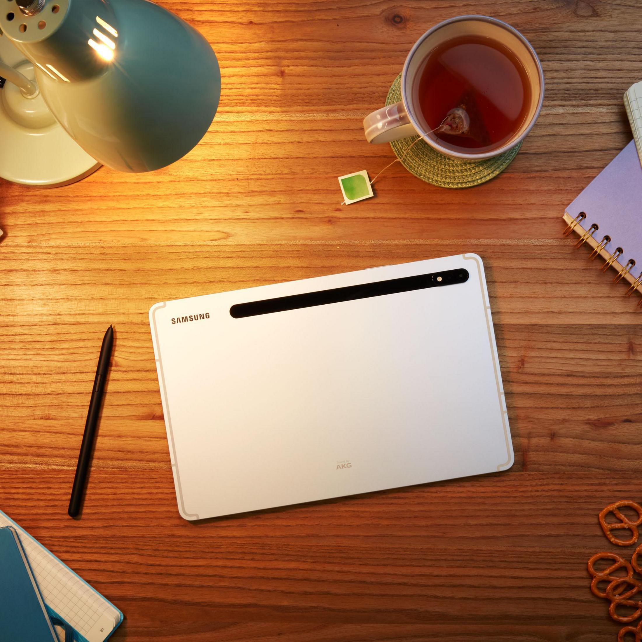 Tablet, Stift, Lampe, Teetasse, Notizblock und Brezeln auf einem Holztisch.