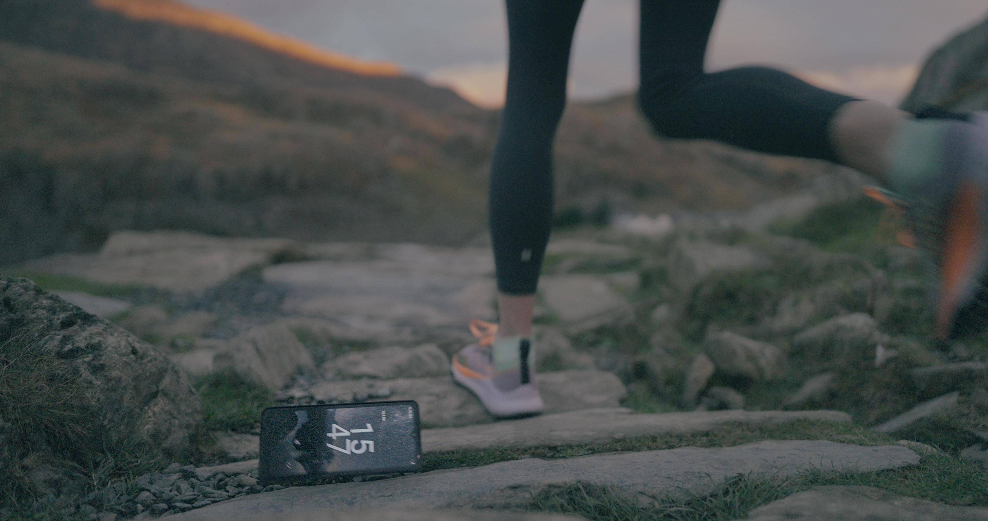 Beine eines Läufers und ein Telefon mit Zeitangabe. Der Läufer auf einem steinigen Weg, Berge im Hintergrund.