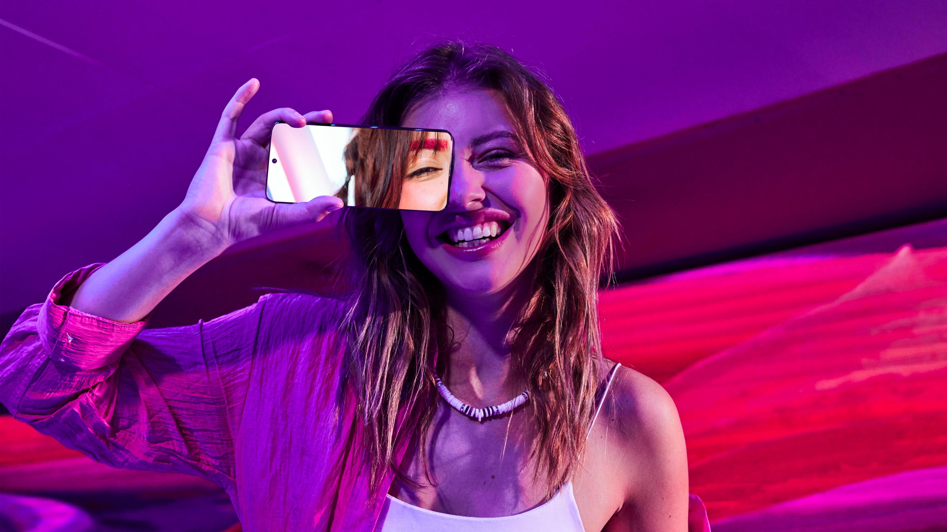 Kobieta trzymająca telefon przed twarzą, ekran odbija jej twarz, na różowo-fioletowym tle.