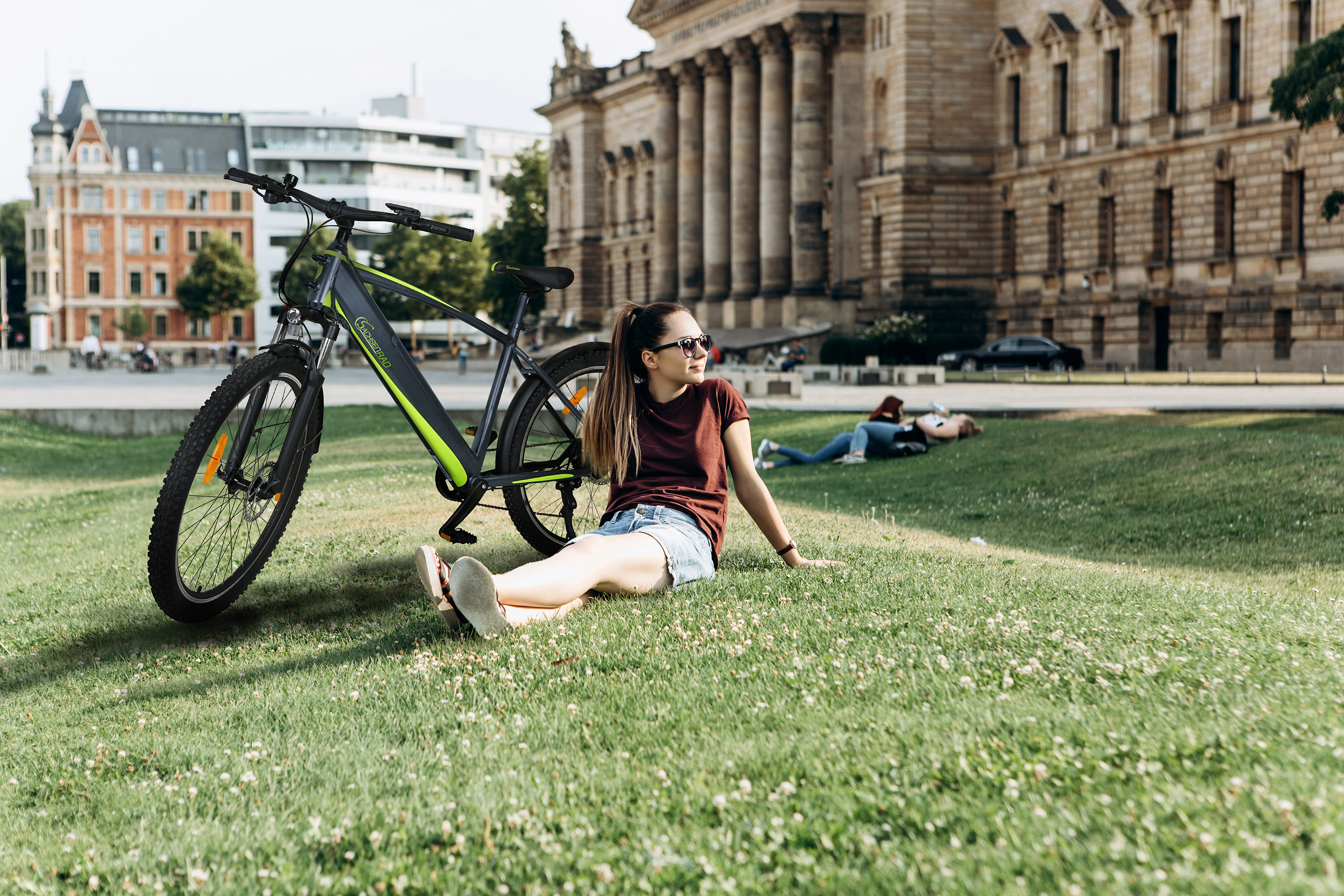 Frau sitzt neben einem Fahrrad auf Gras, Gebäude im Hintergrund.