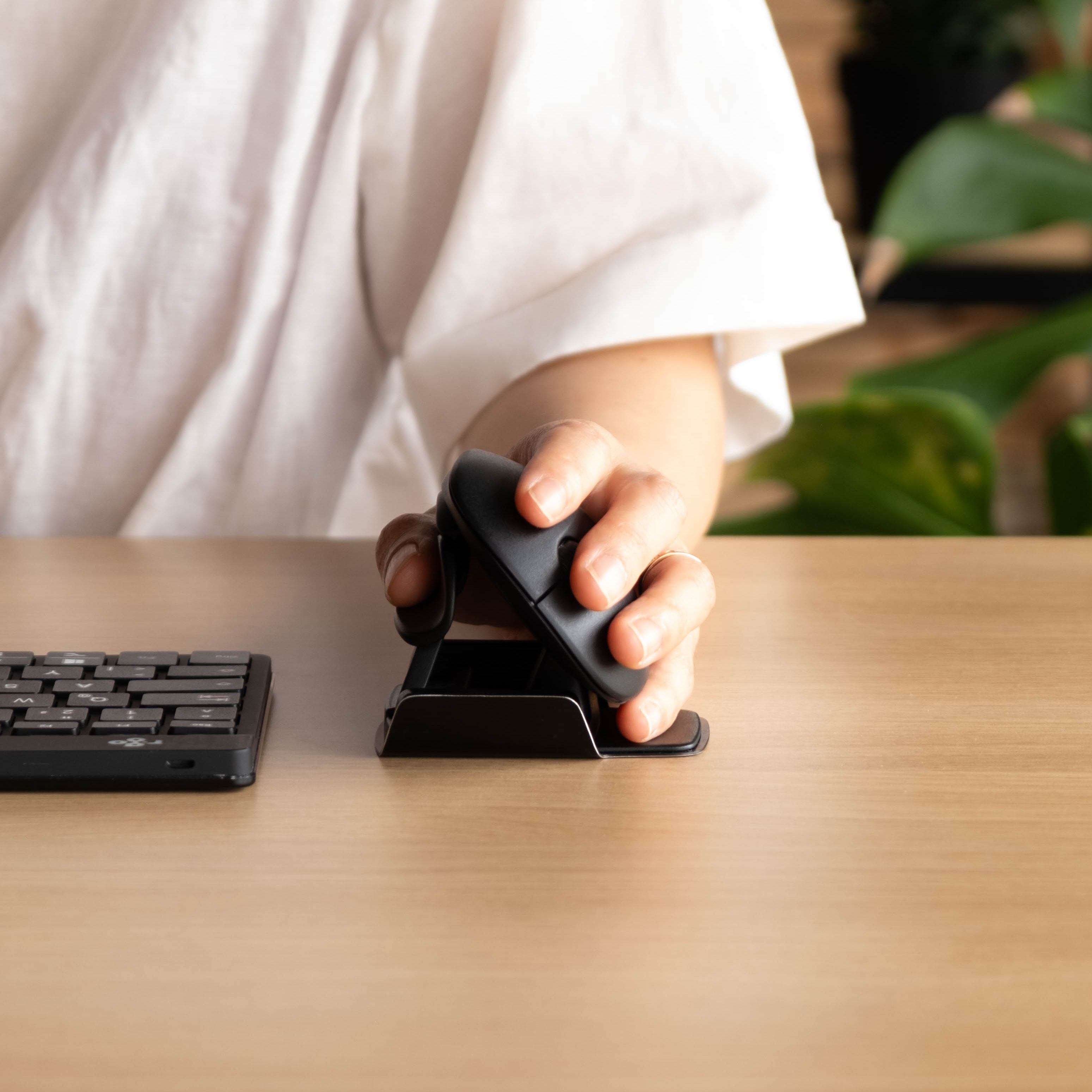 Hand van een persoon die een zwarte ergonomische muis gebruikt op een houten bureau, met een toetsenbord op de achtergrond.