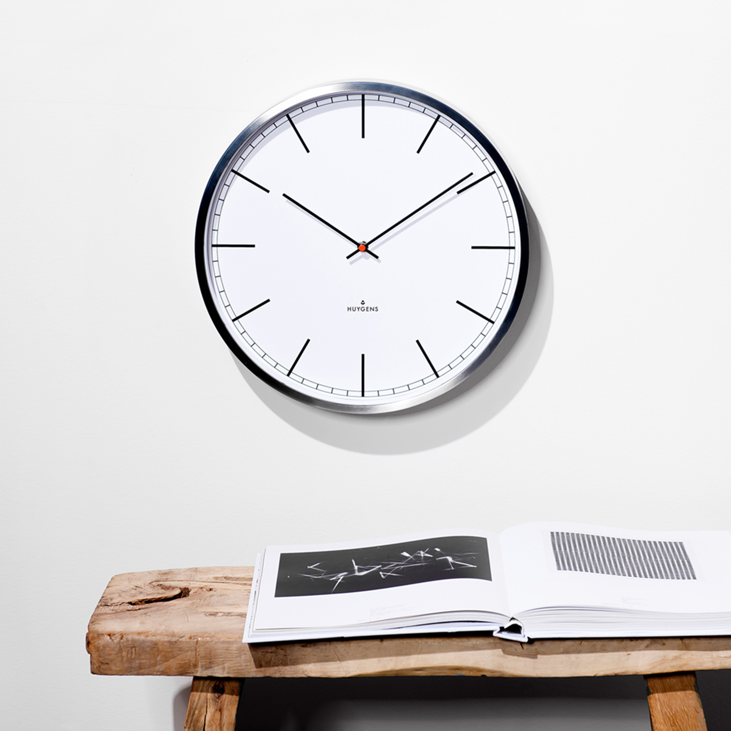 Weiße Wanduhr mit schwarzen Zeigern und Markierungen. Ein Buch liegt darunter auf einer Bank.