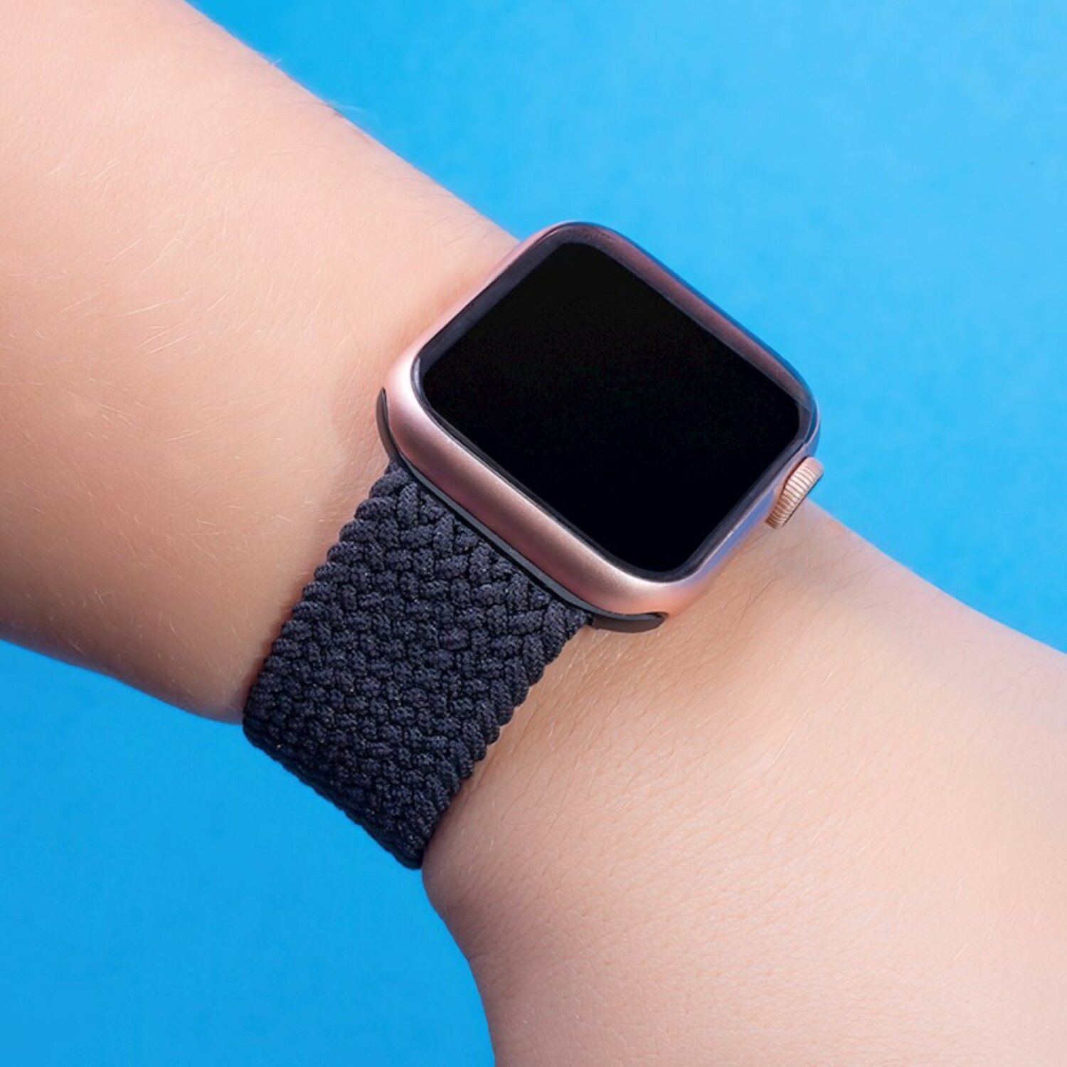 Handgelenk mit roségoldener Apple Watch und dunklem gewebten Armband, vor blauem Hintergrund.