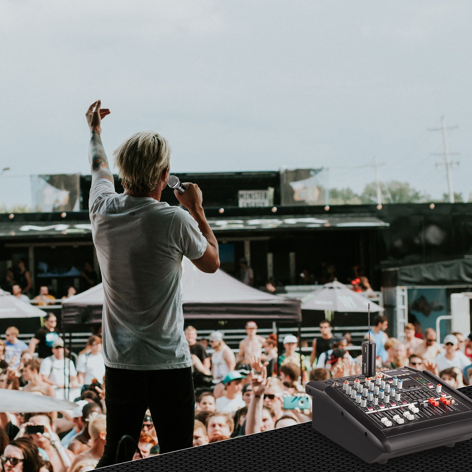Piosenkarz występuje na scenie, podnosząc rękę. Mikser dźwięku jest na pierwszym planie, a tłum w tle.