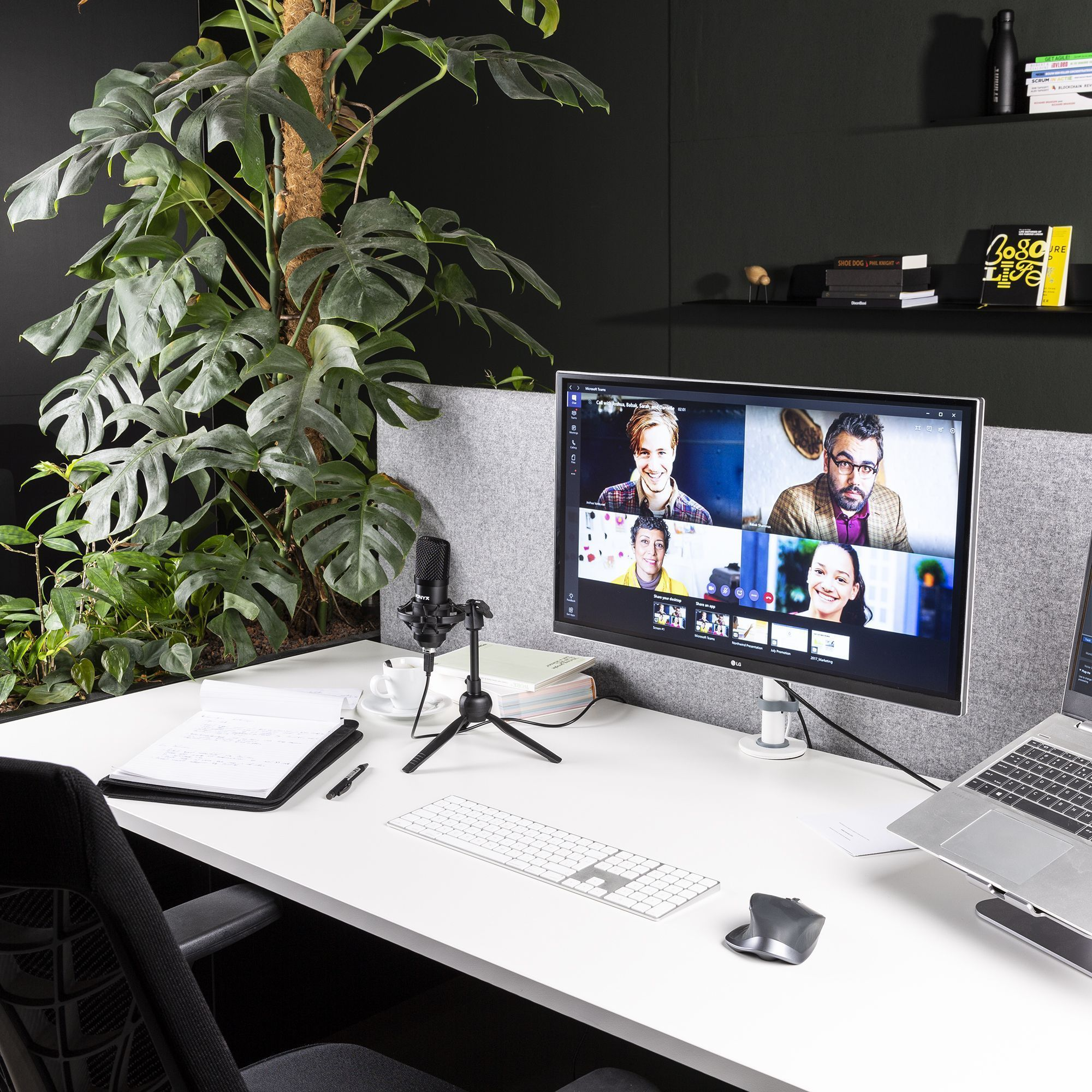 Configuración de oficina con monitor mostrando videollamada. Planta, portátil y micrófono.