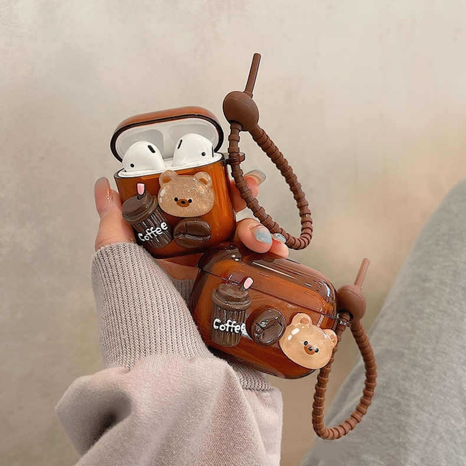 Zwei braune AirPod-Hüllen mit Bären- und Kaffee-Dekorationen, gehalten von Händen vor neutralem Hintergrund.