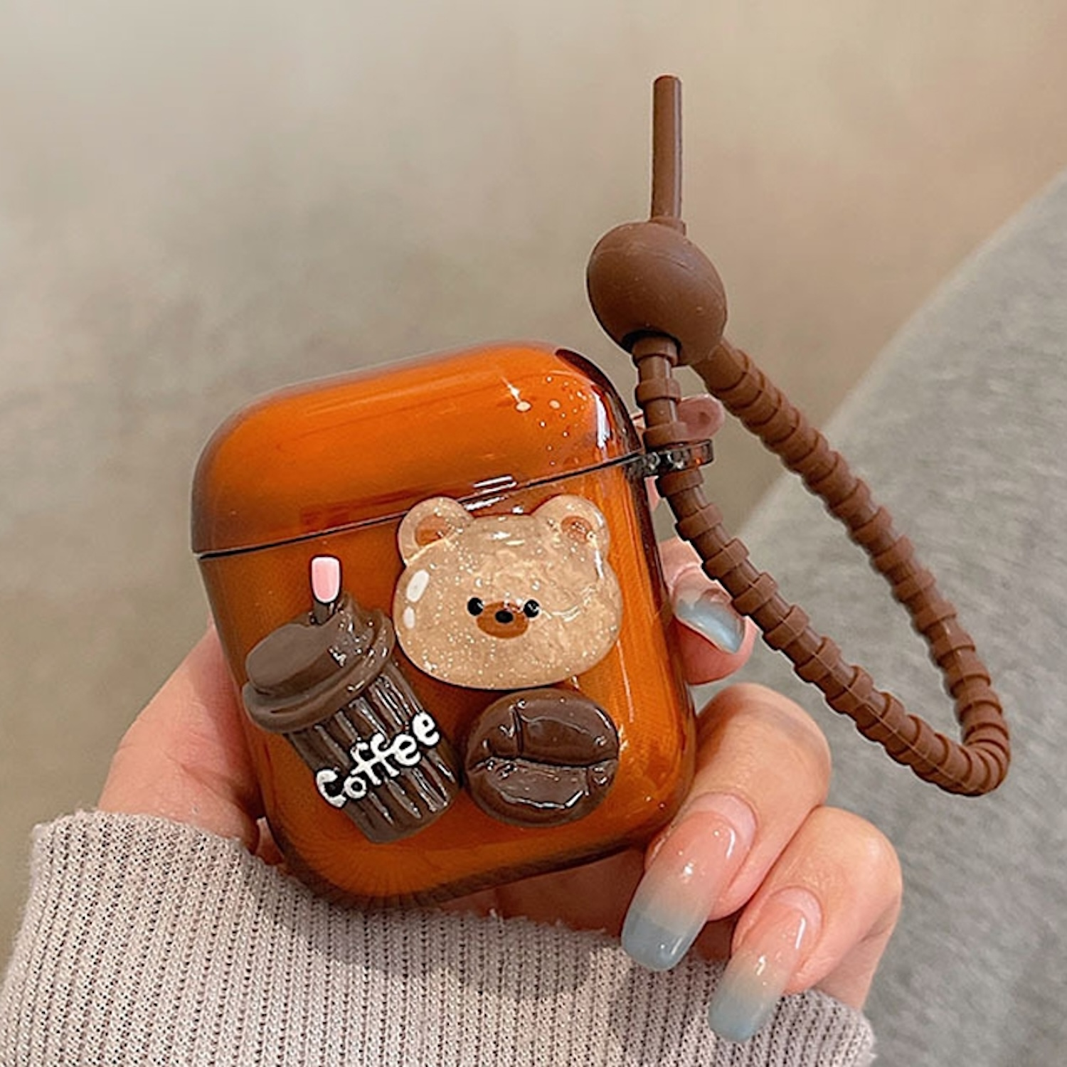 Orange AirPod-Hülle mit Bären- und Kaffee-Dekorationen. Brauner Riemen. Hand hält. Unscharfer Hintergrund.