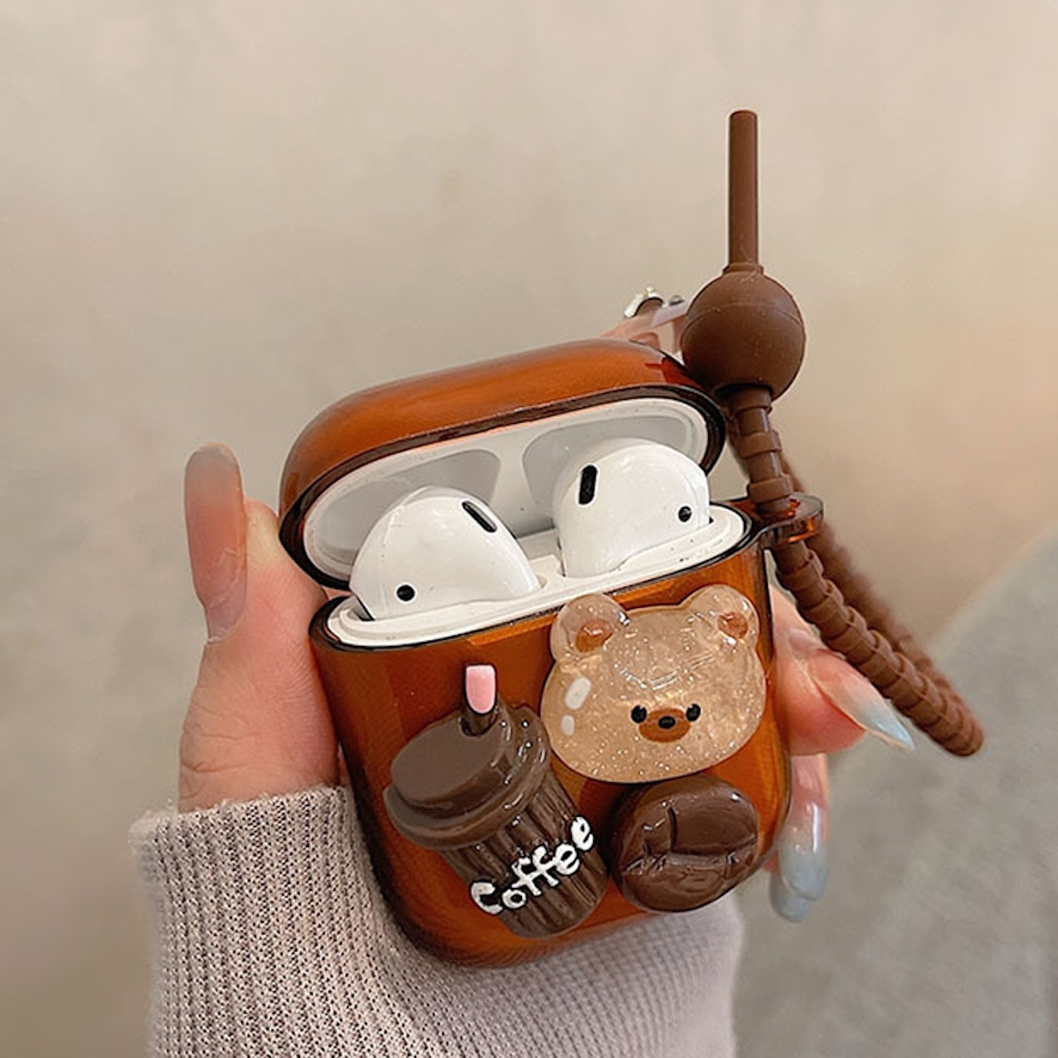 Hand hält eine braune AirPod-Hülle im Kaffeethema mit AirPods darin sowie einem Bären- und Kaffeebohnen-Design.