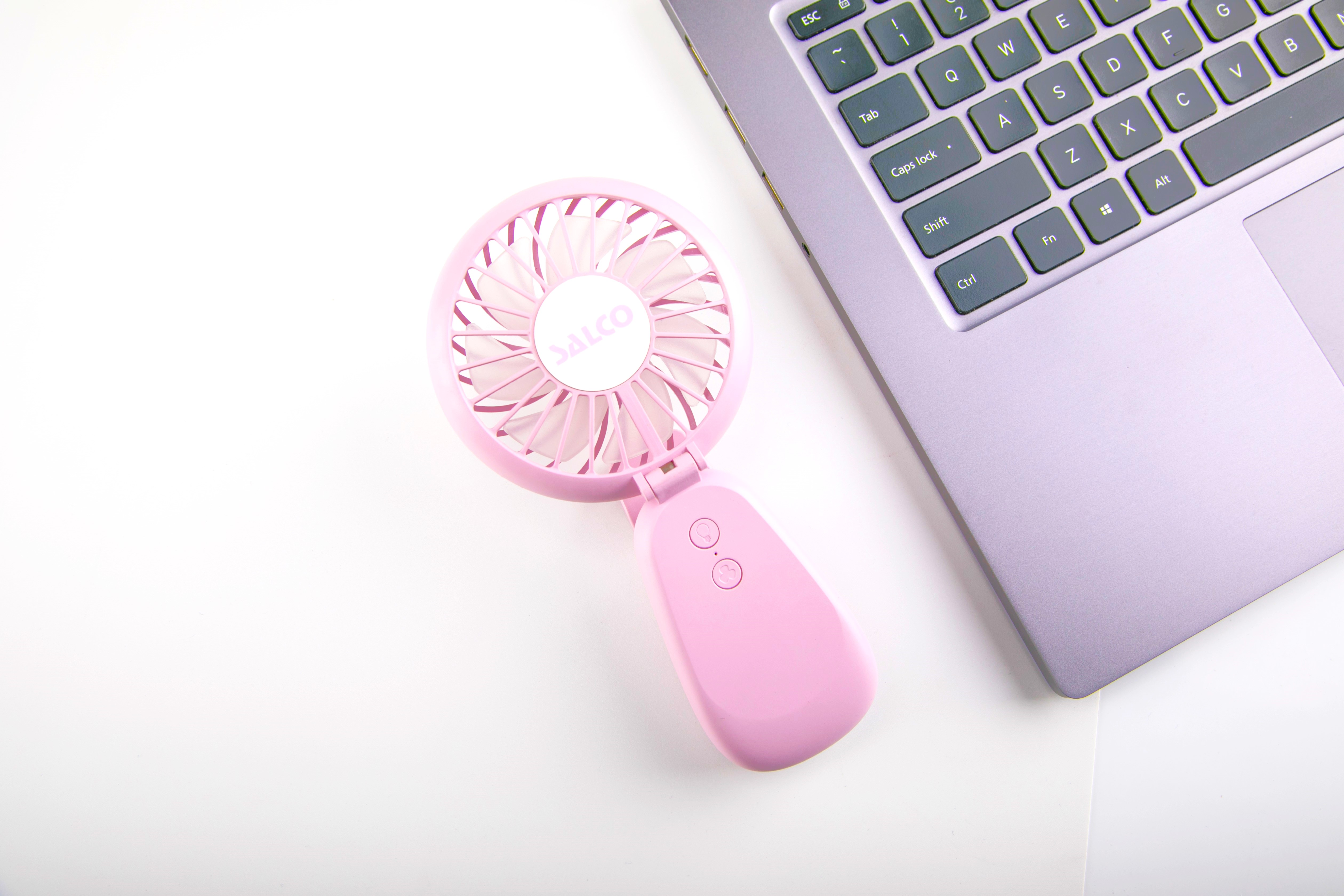 Rosa tragbarer Ventilator neben einem Laptop auf einer weißen Oberfläche.