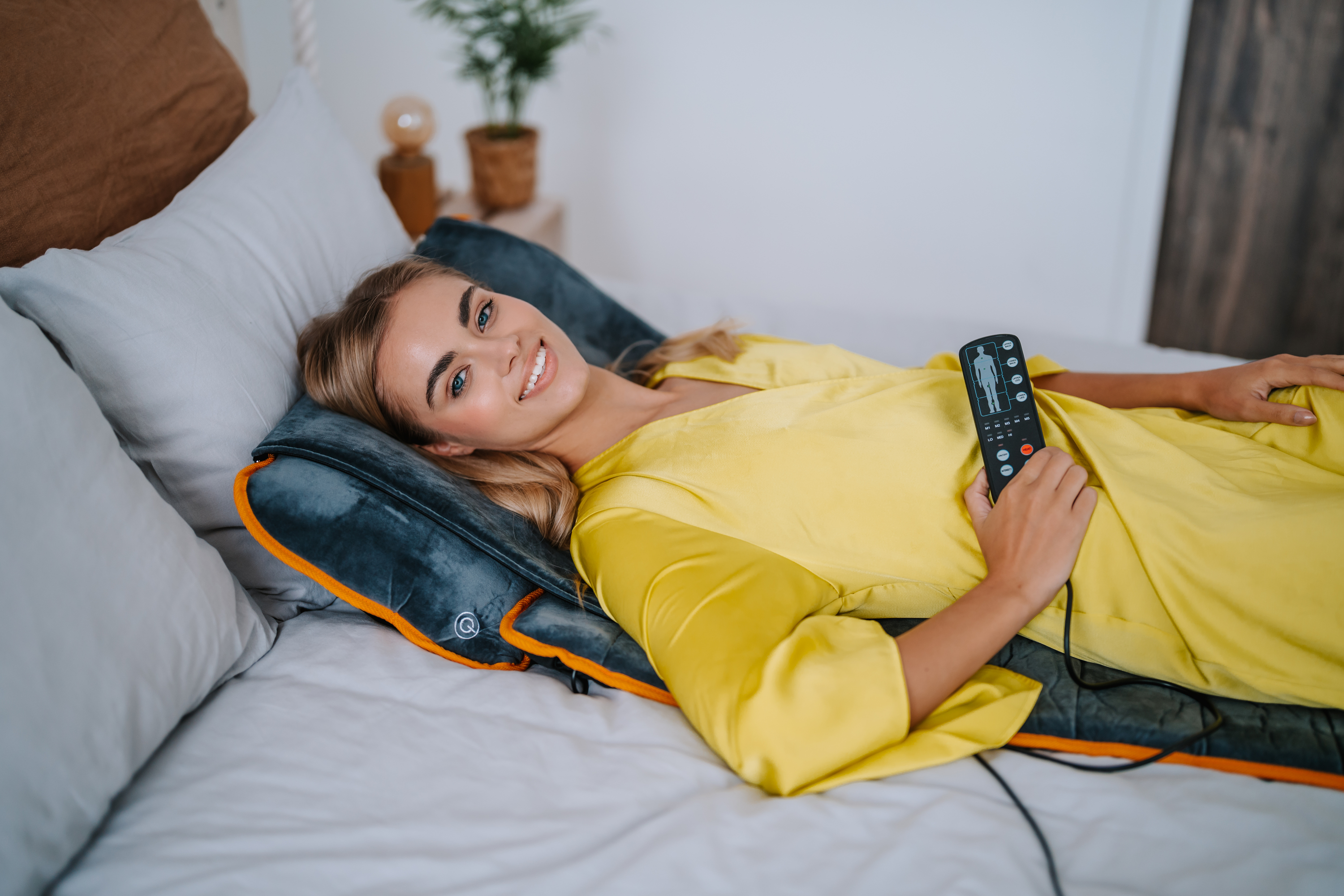 Mujer acostada en una cama con un vestido amarillo, sosteniendo un control remoto. Debajo de ella hay una almohadilla de masaje.