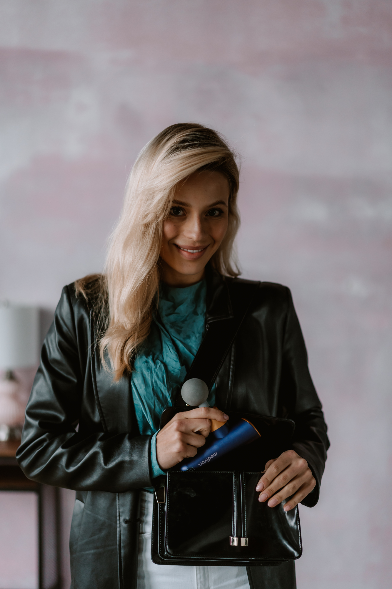 Frau mit blauer Massagepistole, schwarze Jacke, lächelt vor rosa Wand.