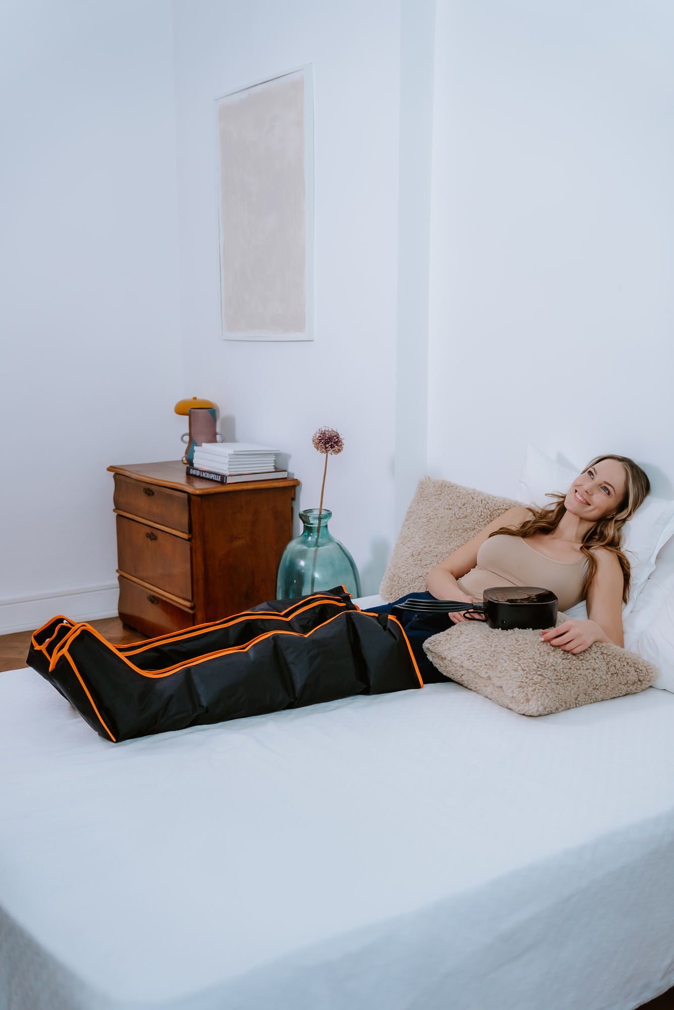 Frau auf dem Bett mit Bein-Erholungsgerät. Sie lächelt, trägt ein Tanktop und ruht auf einem Kissen.