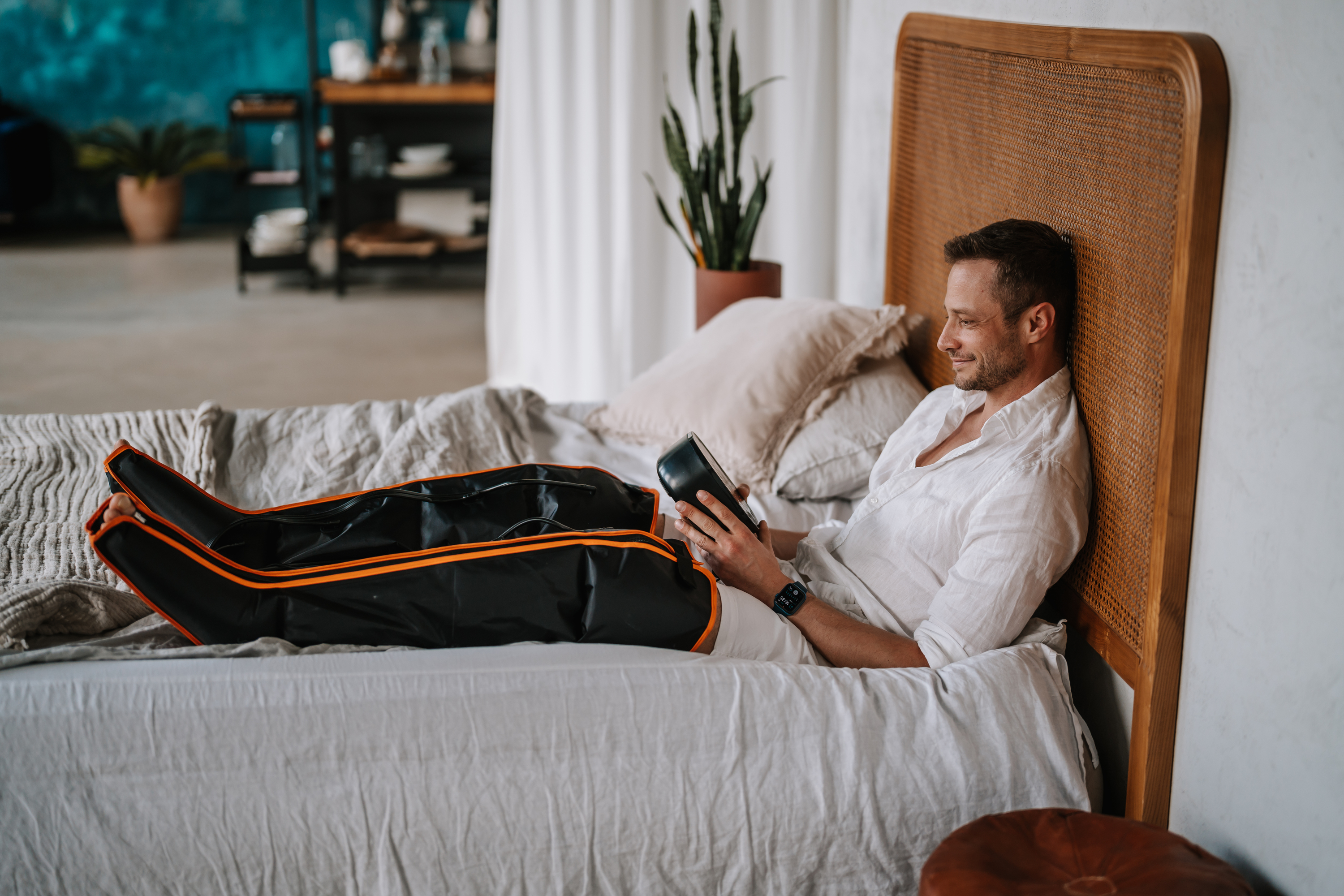 Mann auf Bett mit Beinmassagegerät, liest Tablet.