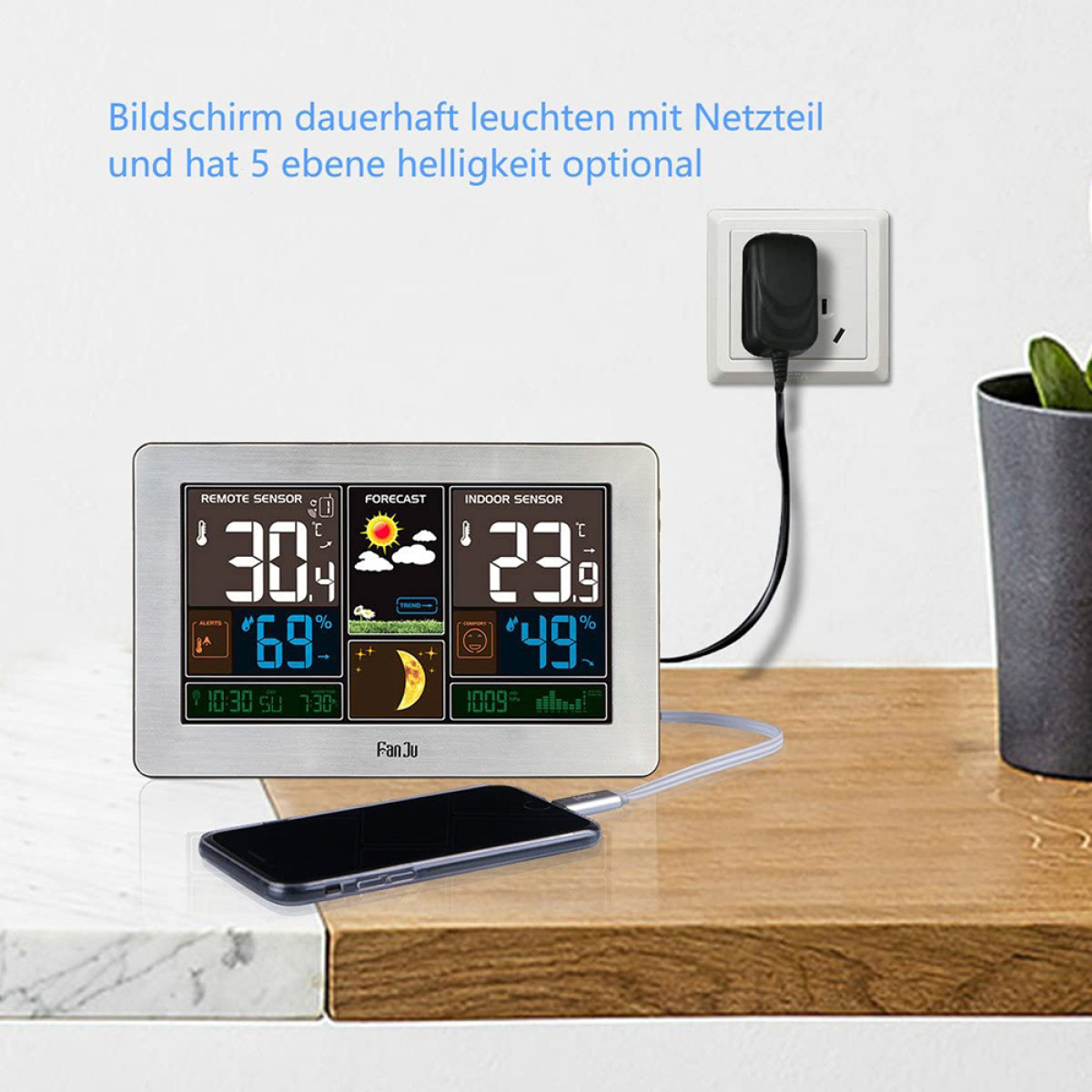 Digitale Wetterstation zeigt Temperatur und Luftfeuchtigkeit. Ein Telefon lädt auf einer Holzoberfläche.