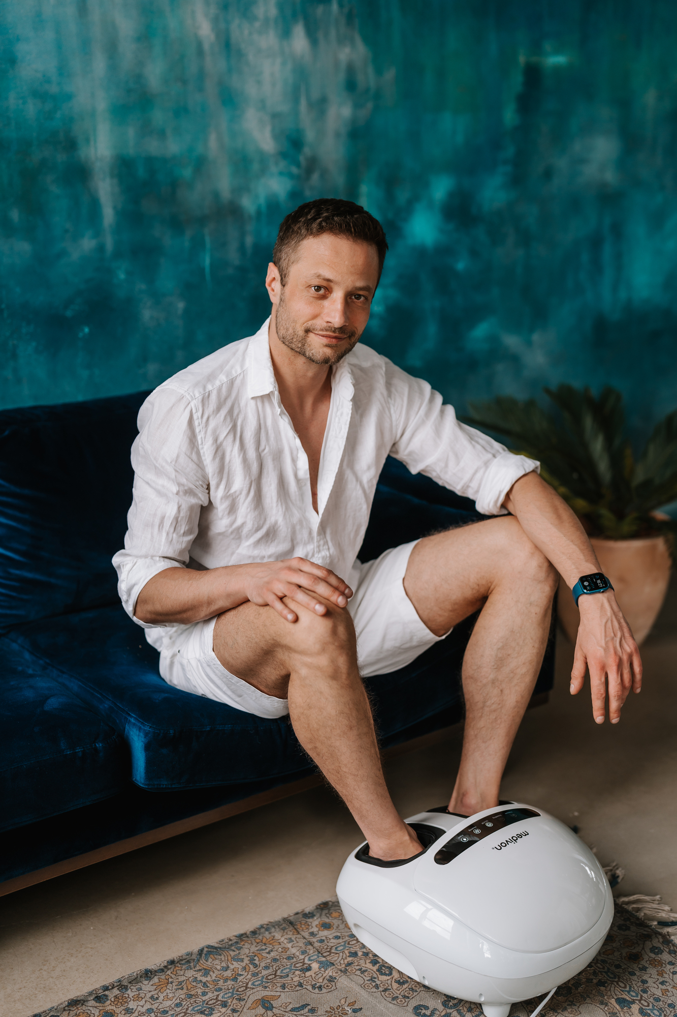 Mann sitzt auf blauem Sofa und benutzt Fußmassagegerät. Er lächelt.