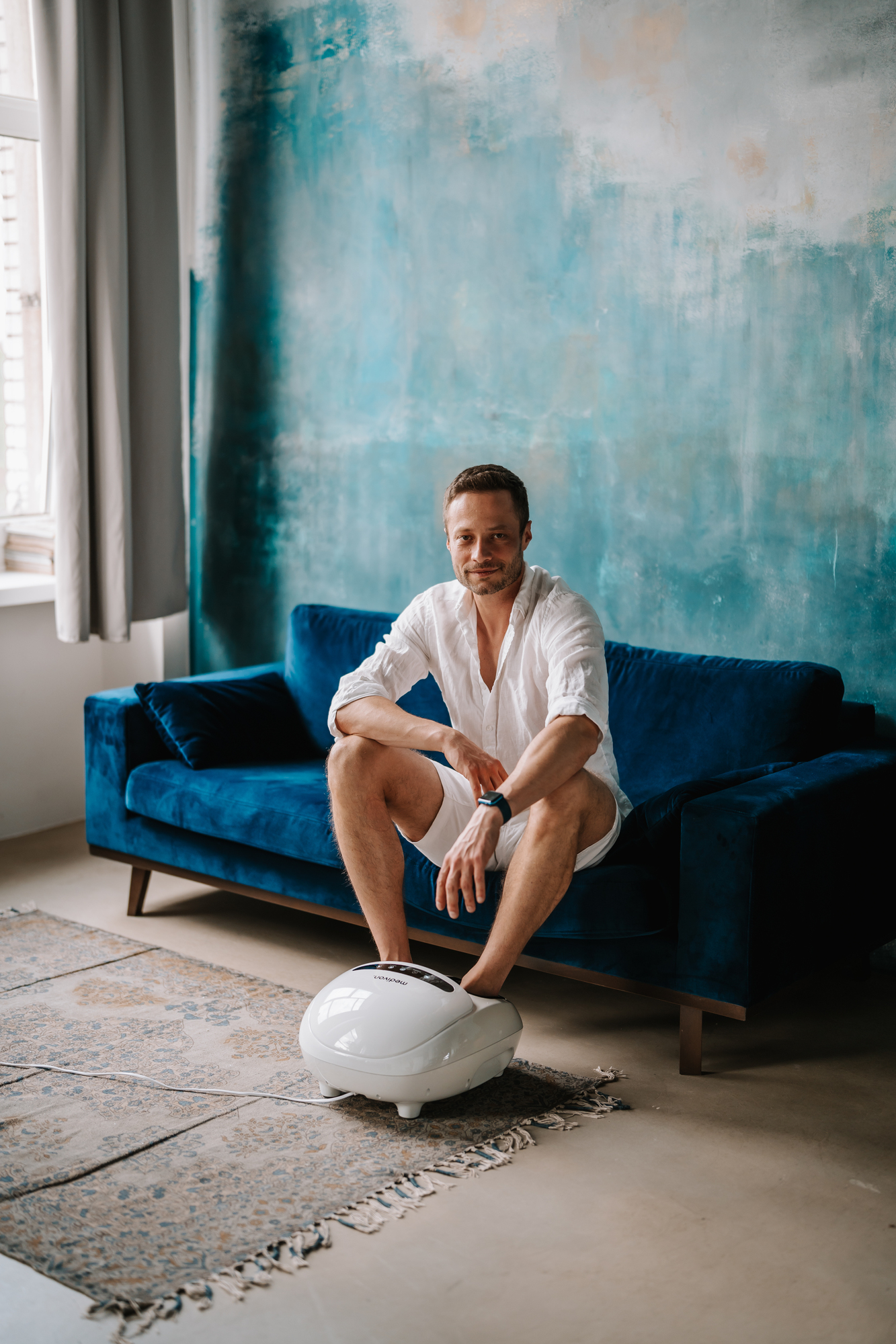 Mann sitzt auf blauem Sofa, Fußmassagegerät an den Füßen, blauer Wandhintergrund.