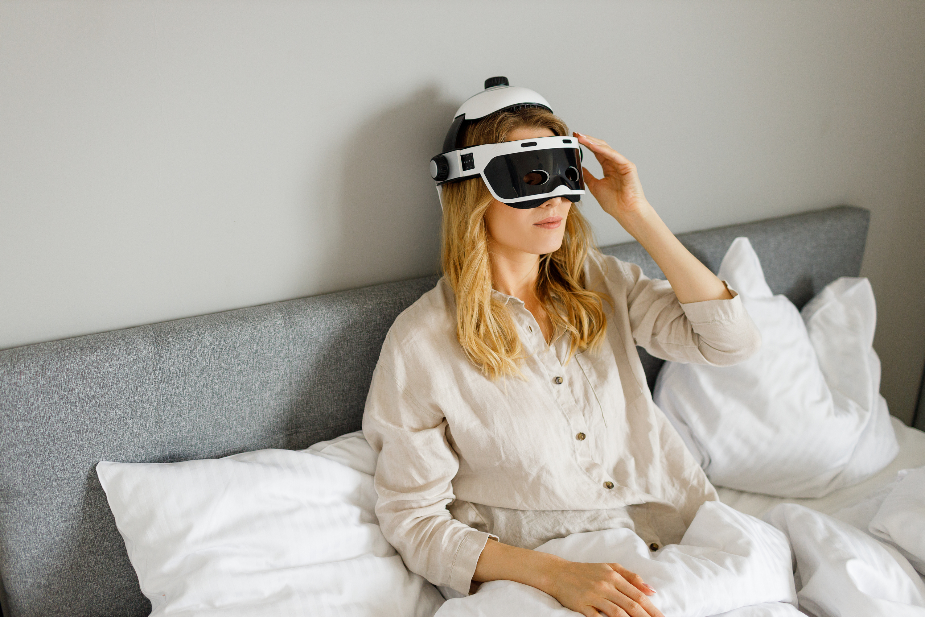 Eine Frau mit VR-Headset sitzt im Bett und berührt es mit der Hand.