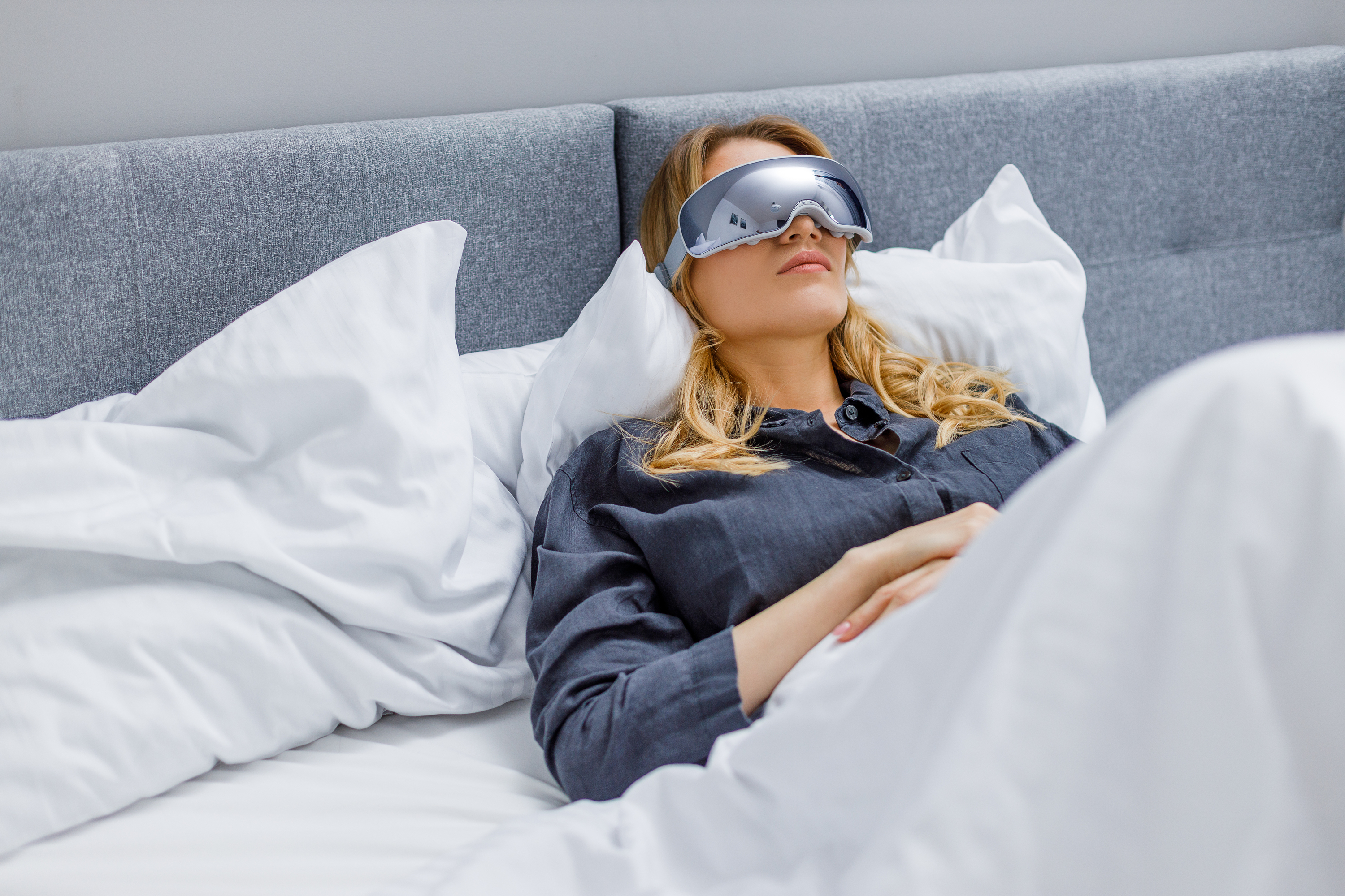 Eine schlafende Frau im Bett mit Schlafmaske. Sie liegt unter einer weißen Bettdecke und auf einem grauen Kopfteil.