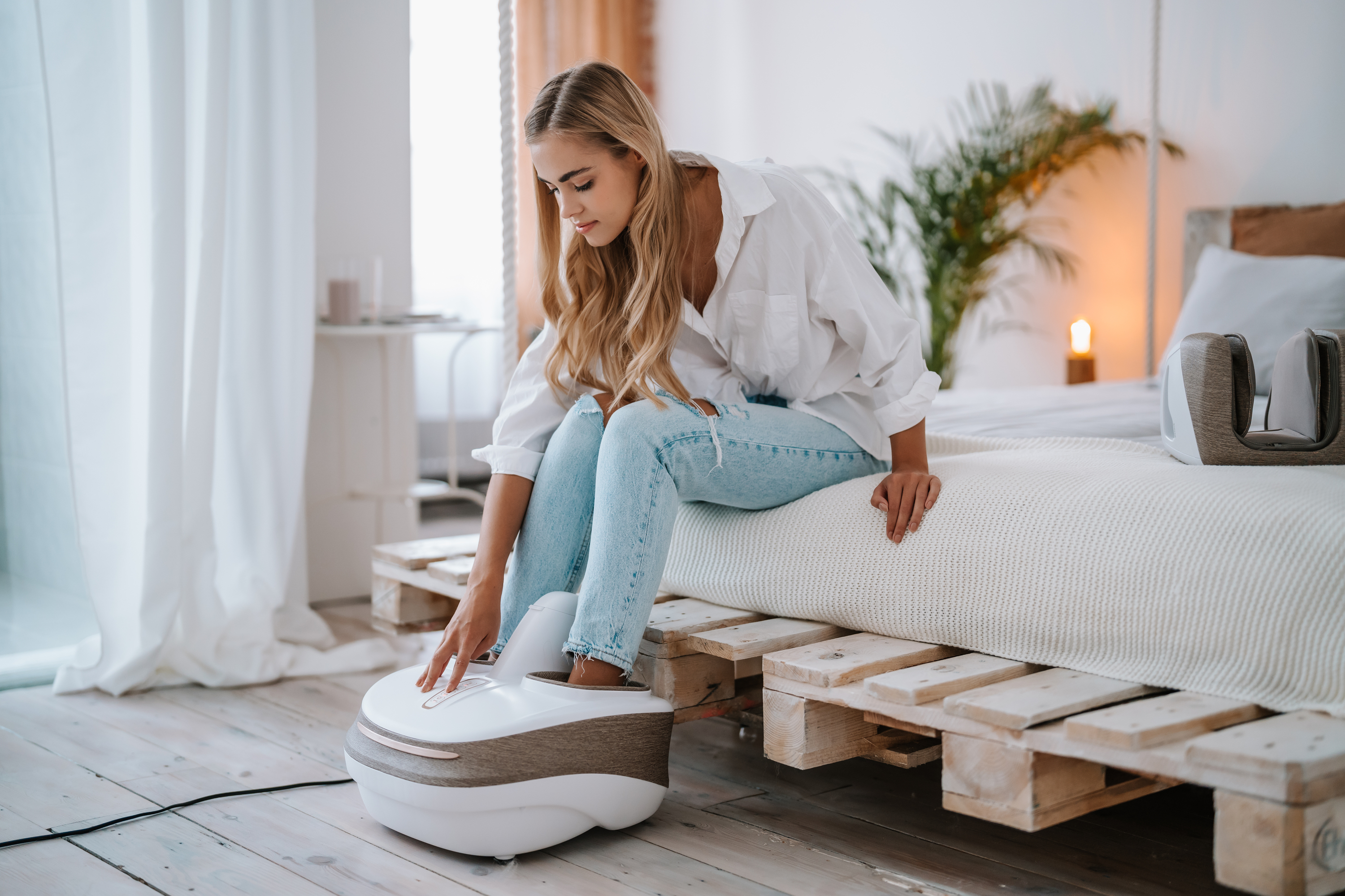 Eine Frau in Jeans und weißem Hemd benutzt ein Fußmassagegerät auf einem Bett mit Holzpalettenbasis.