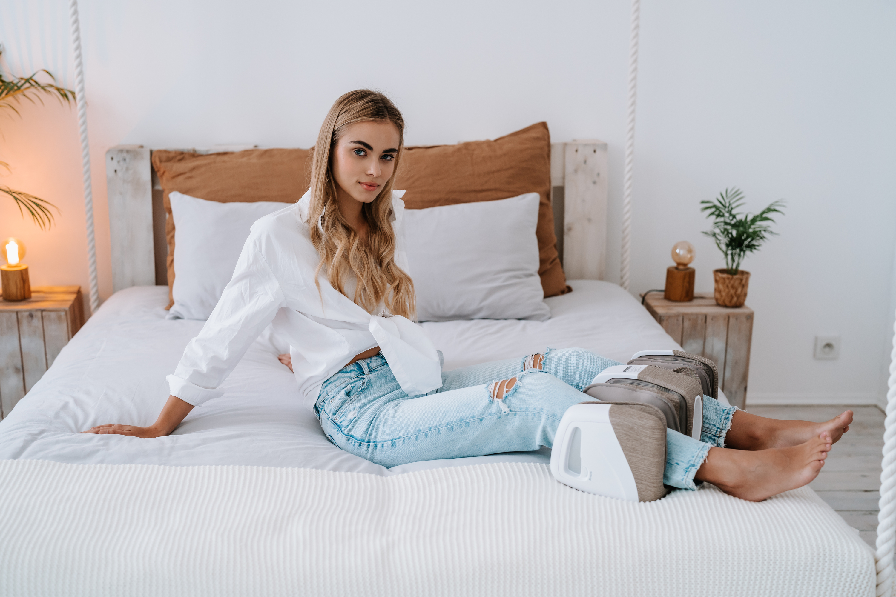 Frau in Jeans und weißem Hemd sitzt auf dem Bett. Sie benutzt Beinmassagegeräte.
